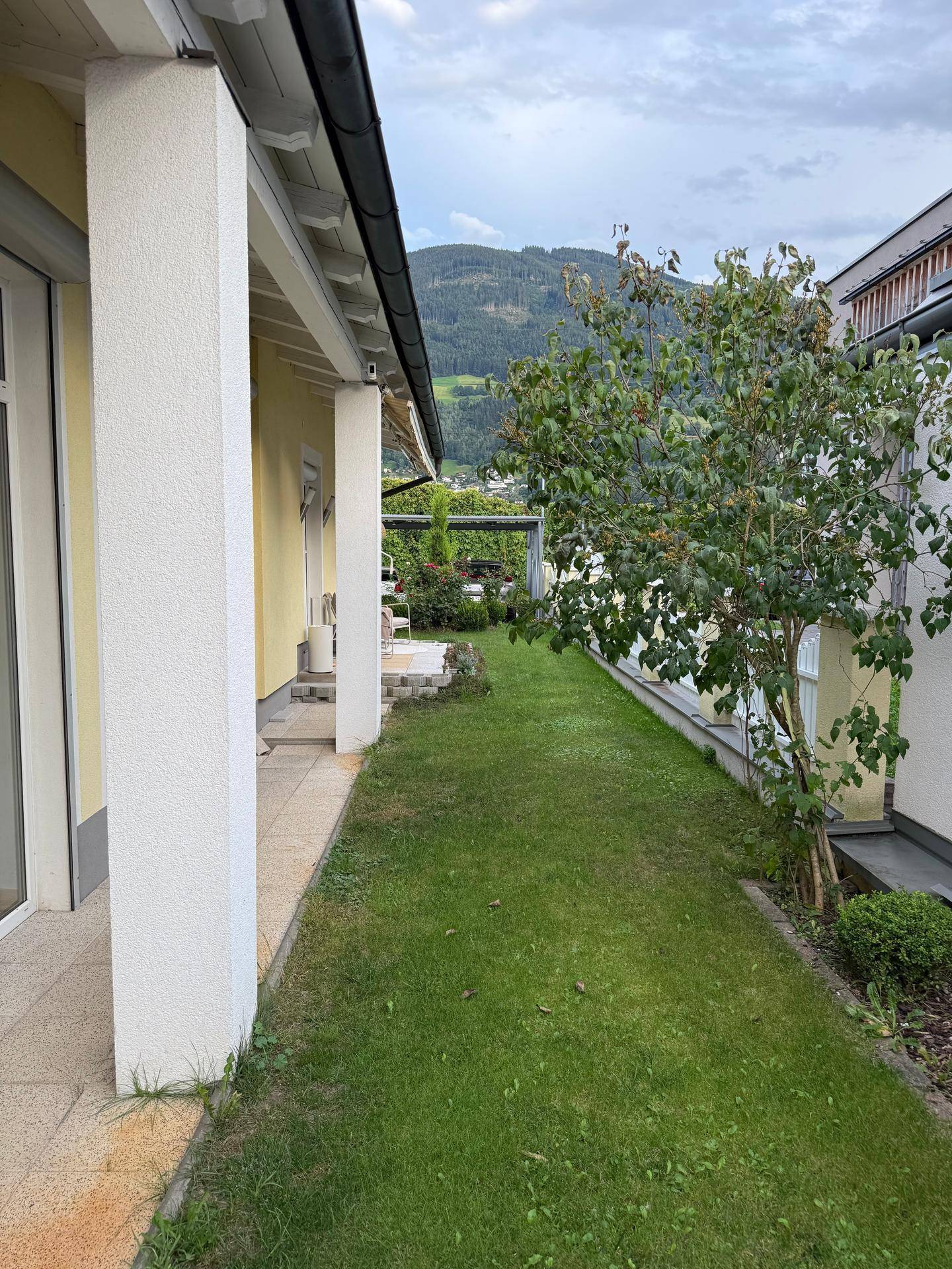 Seitlicher Gartenbereich mit Rasenfläche und Zugang zur überdachten Terrasse, umgeben von Natur.