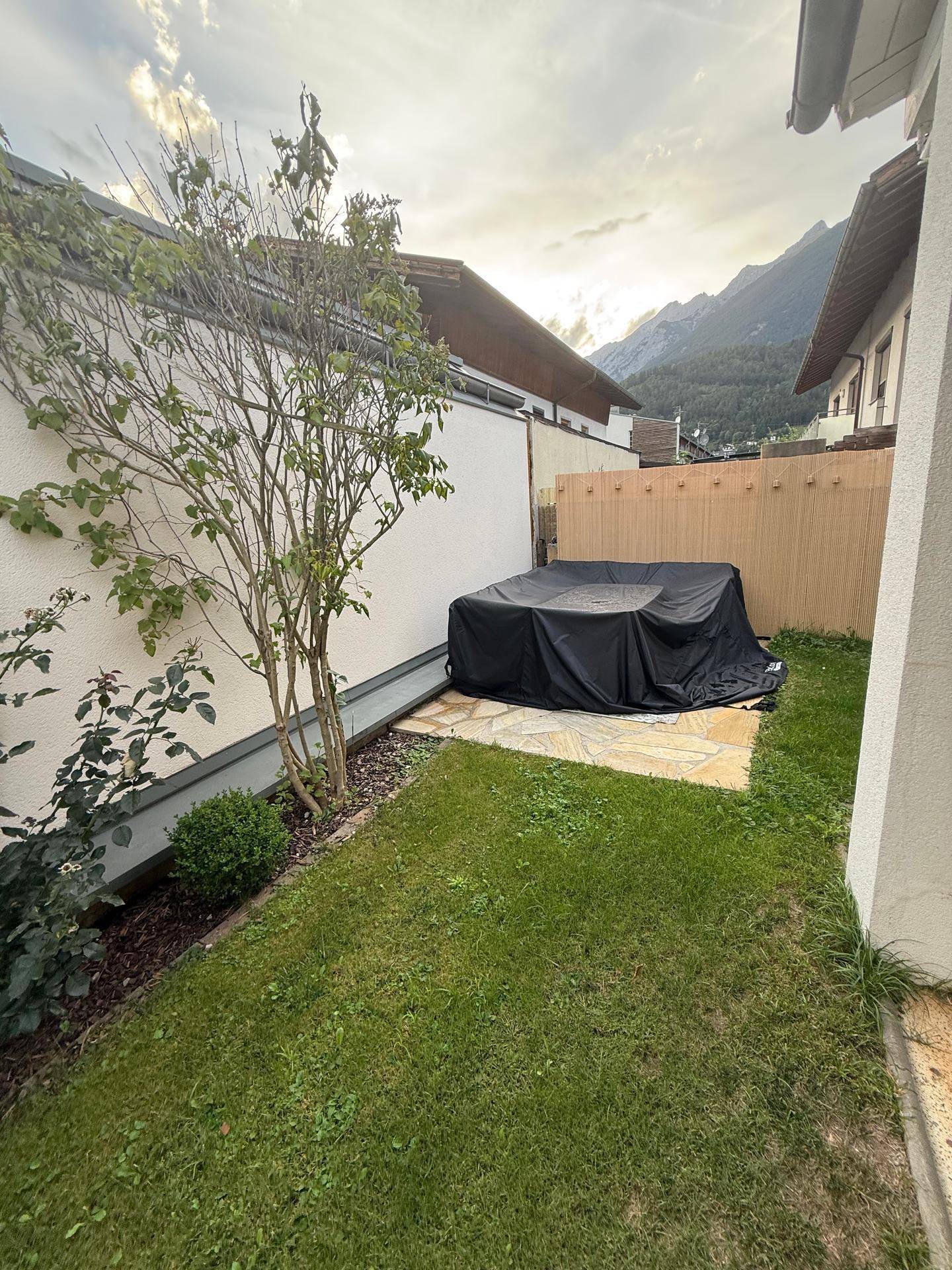Kleiner, gepflegter Gartenbereich mit Rasen, Baum und überdachten Gartenmöbeln.