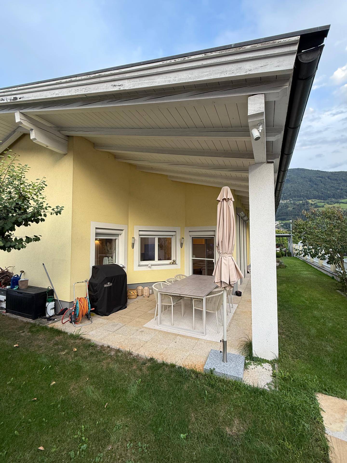 Überdachte Terrasse mit Essbereich und Grill, umgeben von Rasen und mit Bergblick.