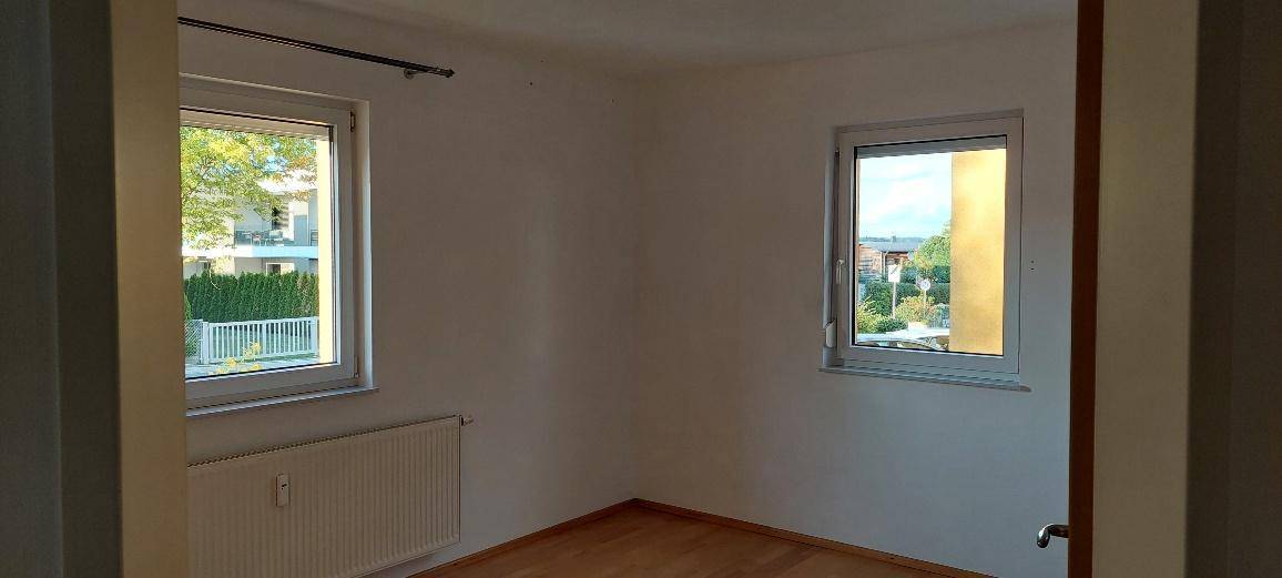 Helles Schlafzimmer mit Holzboden, Heizkörper und zwei Fenstern mit Blick ins Grüne.