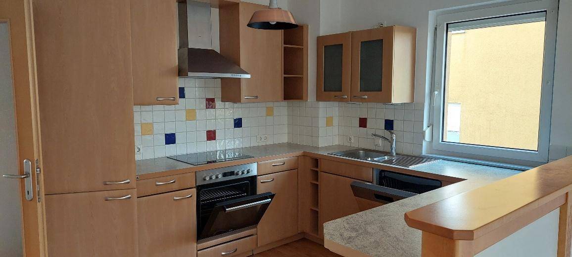 Funktionale Küche mit Holzschränken, Fliesenrückwand und integrierten Geräten, hell durch ein Fenster.