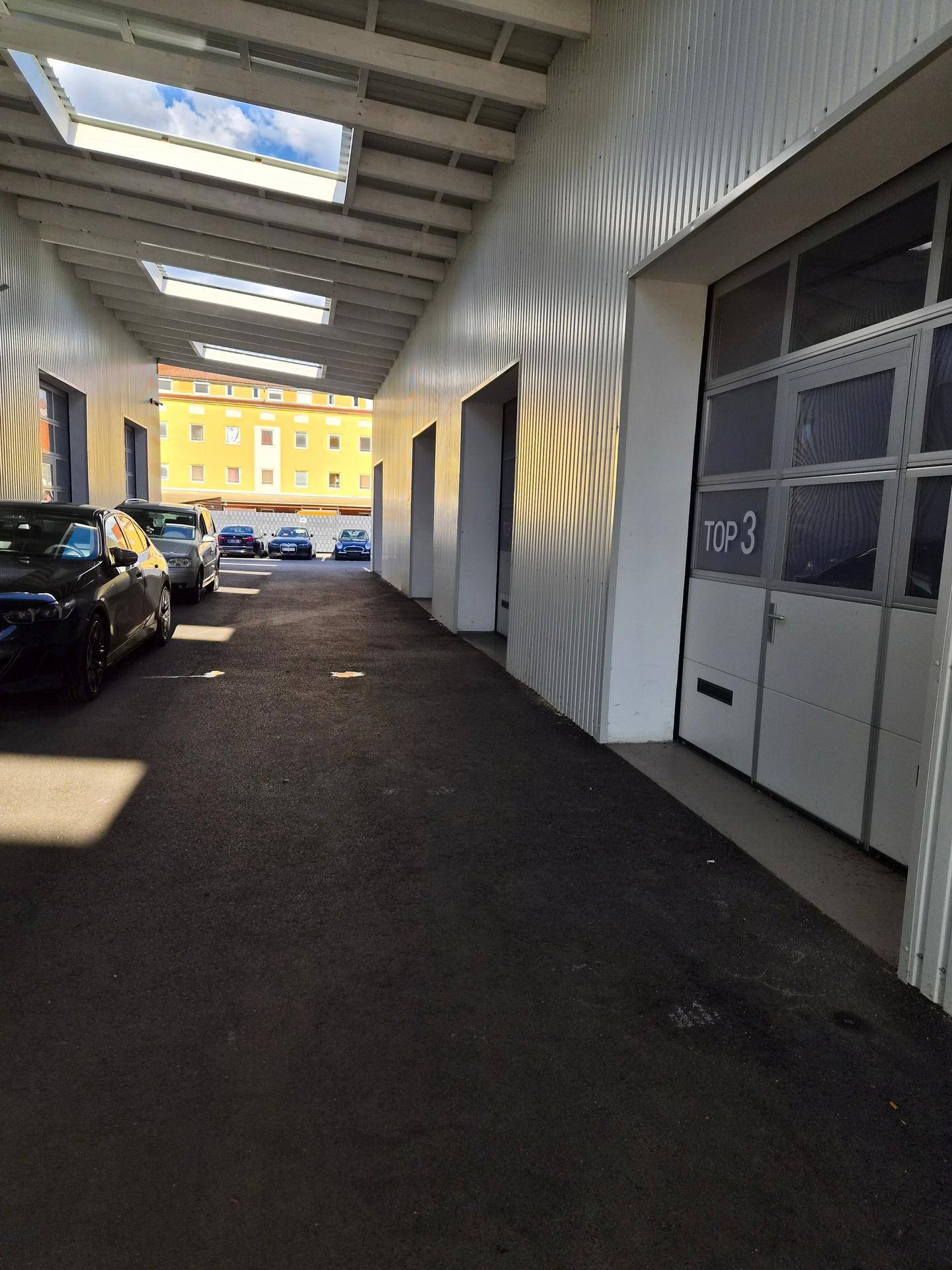 Überdachte Zufahrt mit mehreren Garagentoren und geparkten Autos im Hintergrund, beleuchtet durch Oberlichter im Dach.