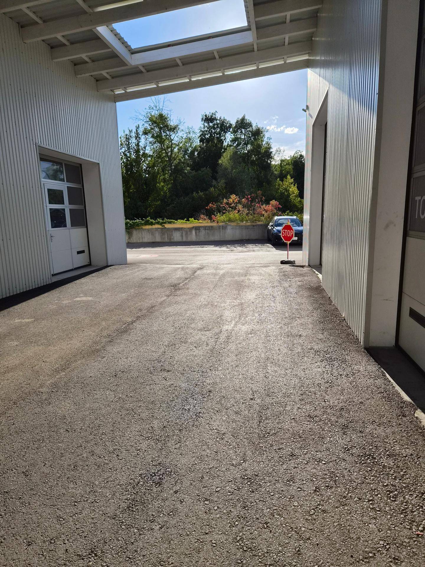 Außenbereich zwischen zwei Gebäuden mit Asphaltboden, einem Stoppschild und Bäumen im Hintergrund unter einem sonnigen Himmel.