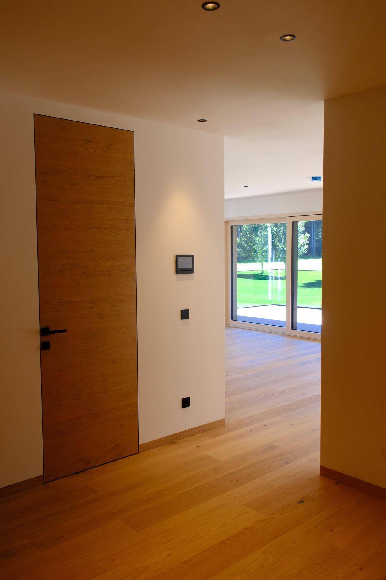Heller Flur mit Holzboden und Blick in einen lichtdurchfluteten Raum mit großen Fenstern.