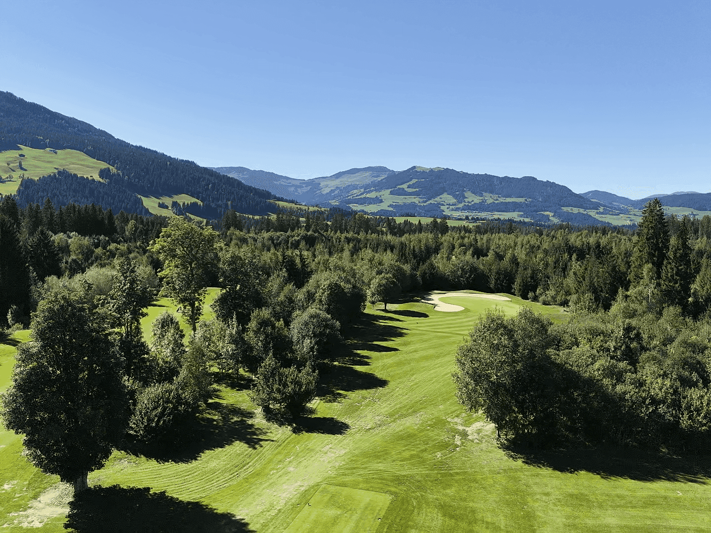Weitläufige Golflandschaft eingebettet in eine grüne Wald- und Bergkulisse unter blauem Himmel.
