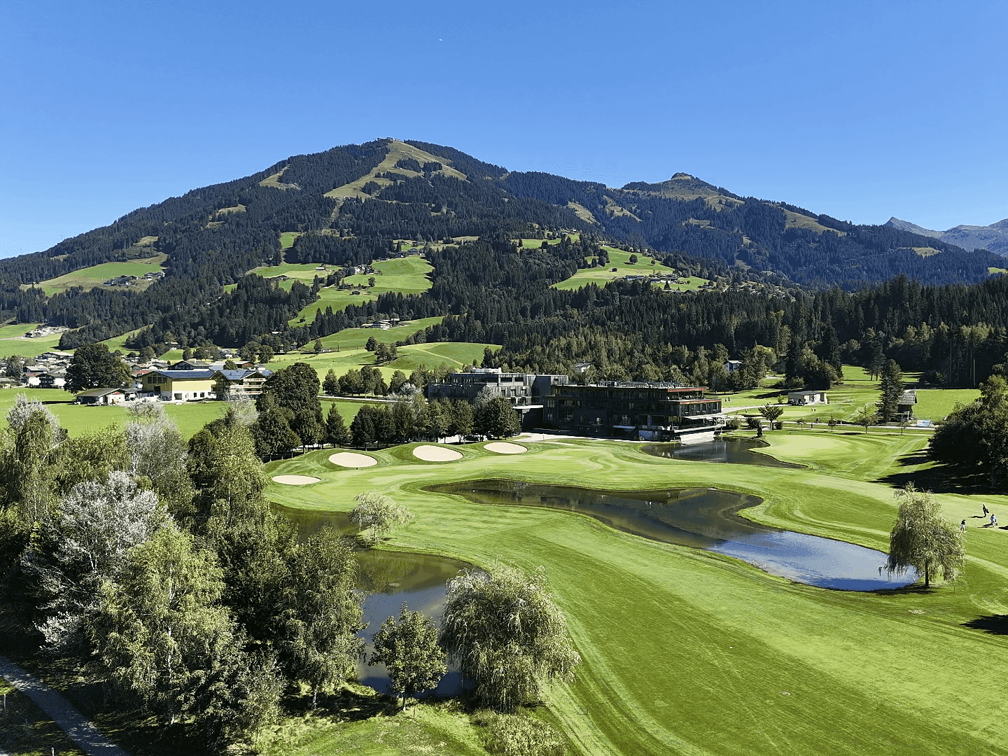 Weitläufige Golfanlage mit Wasserhindernissen, umgeben von Wäldern und einer beeindruckenden Bergkulisse.