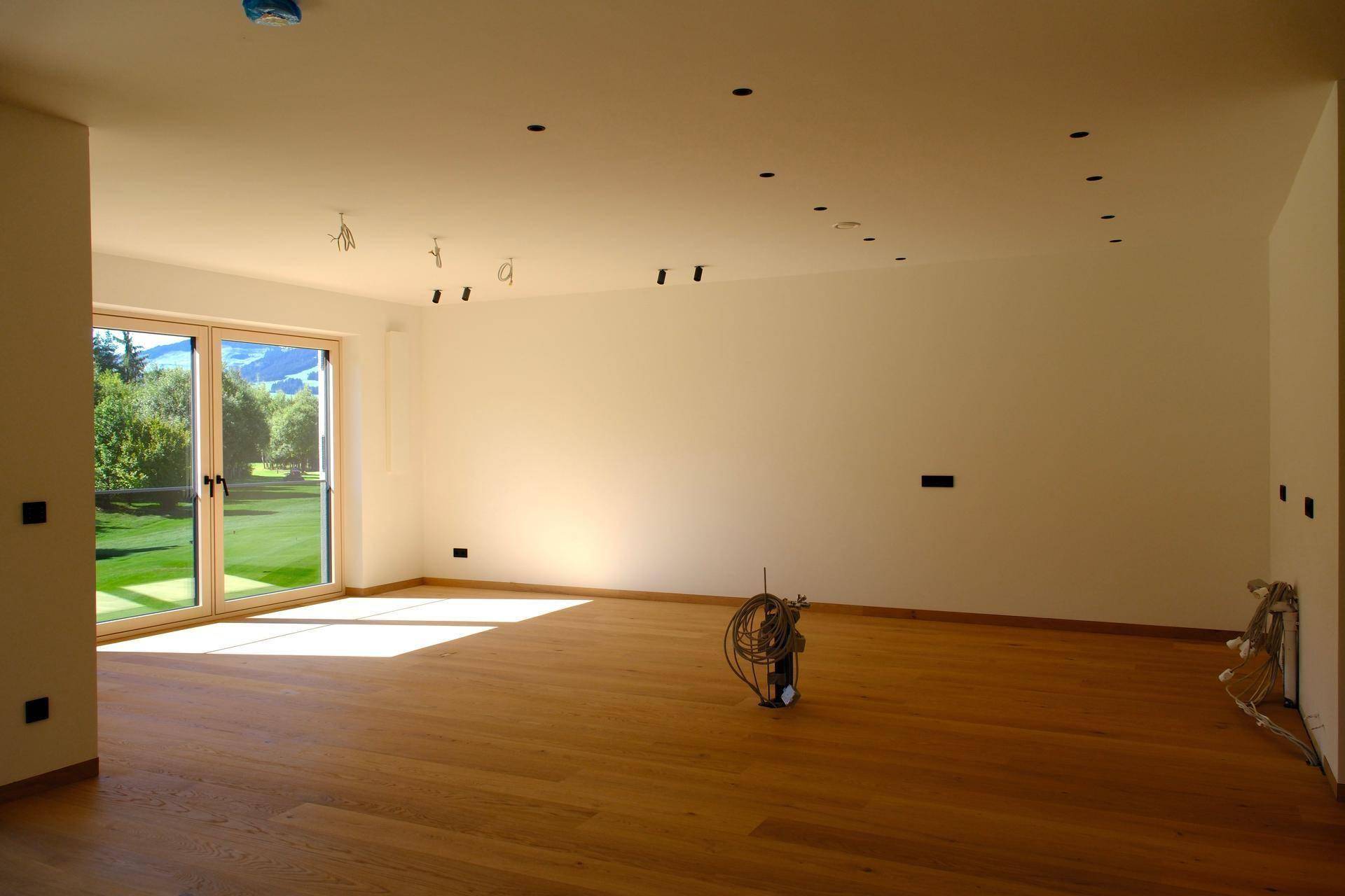 Geräumiges, leeres Wohnzimmer mit Holzboden und großen Fenstern, die viel Tageslicht hereinlassen.