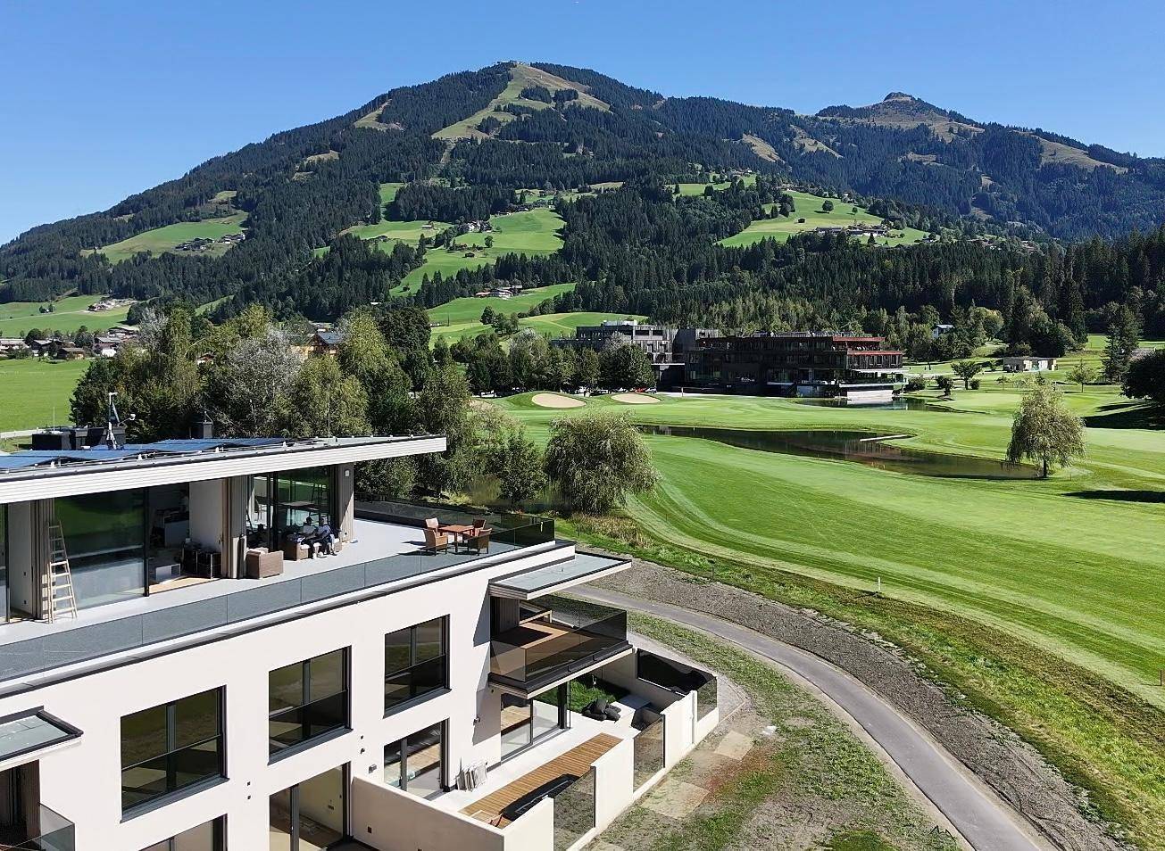 Luftaufnahme eines modernen Gebäudes mit Dachterrasse, umgeben von einem Golfplatz und Bergen.