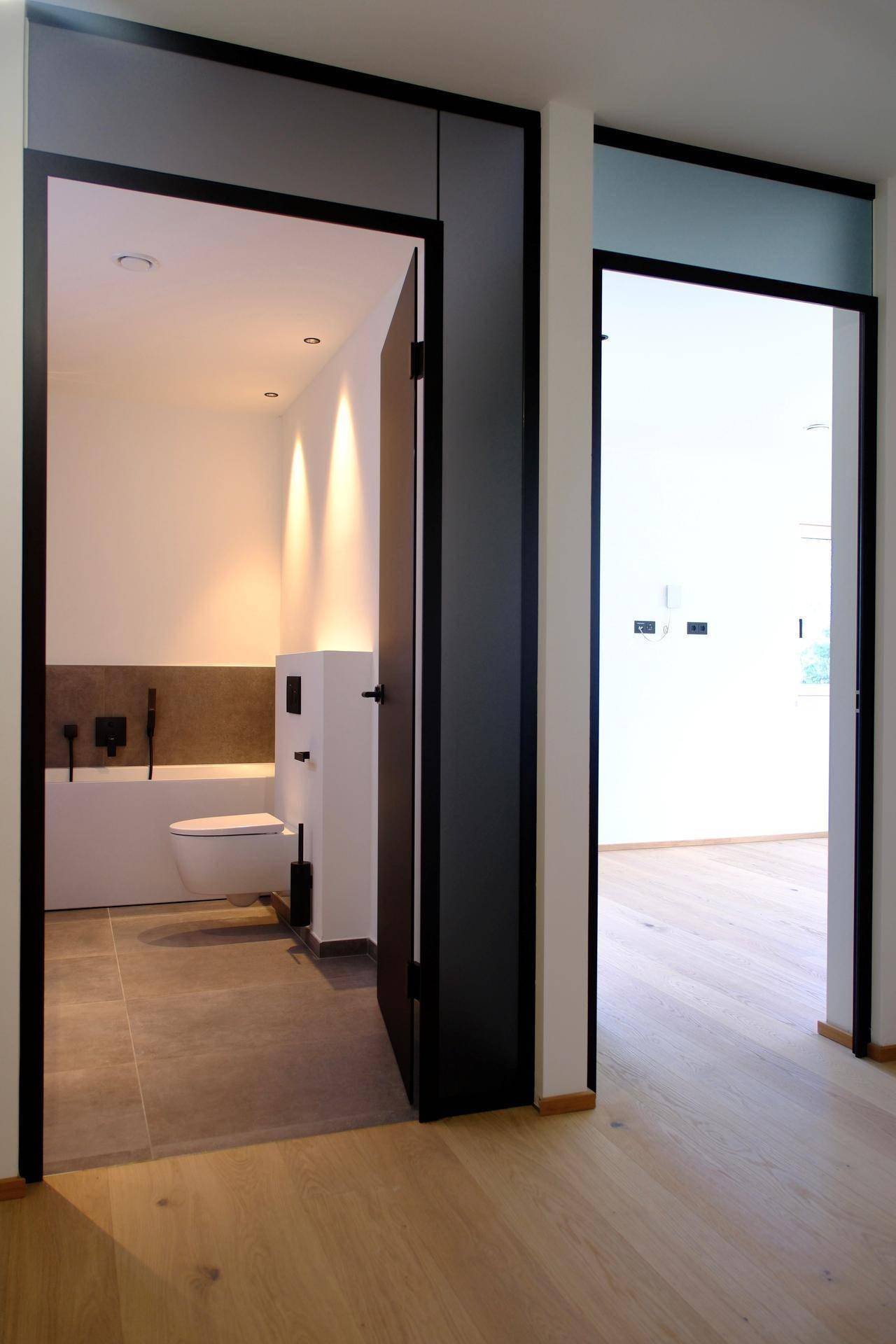 Heller Flur mit Holzboden und Blick in ein modernes Badezimmer mit dunklen Fliesen.