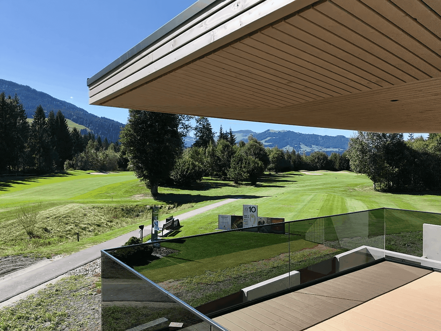 Balkon mit Holzboden und Glasgeländer, bietet einen Panoramablick auf den Golfplatz und die Berge.