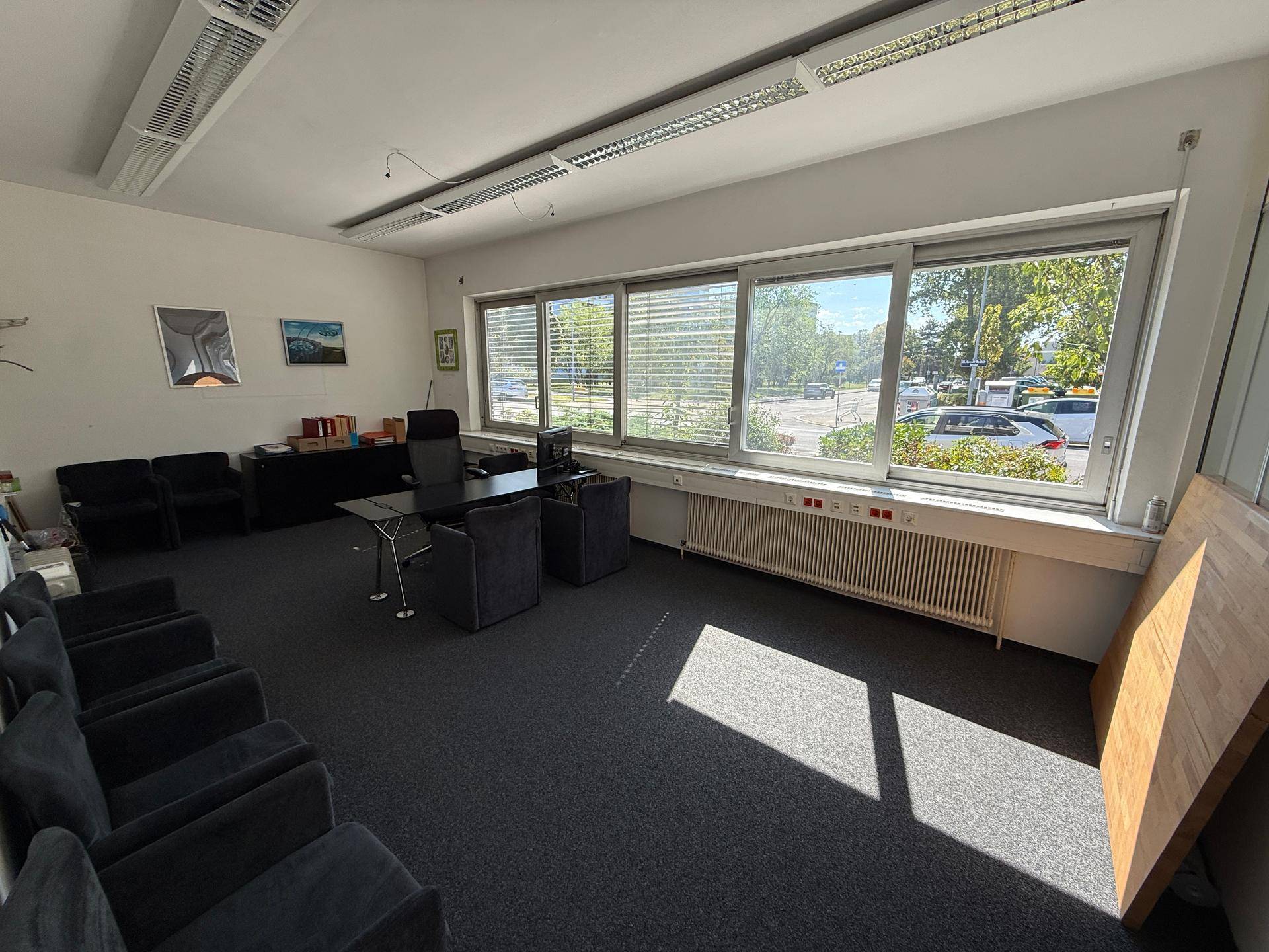 Geräumiges Büro mit großen Fenstern, Teppichboden und mehreren Sitzgelegenheiten, gut beleuchtet durch Tageslicht.