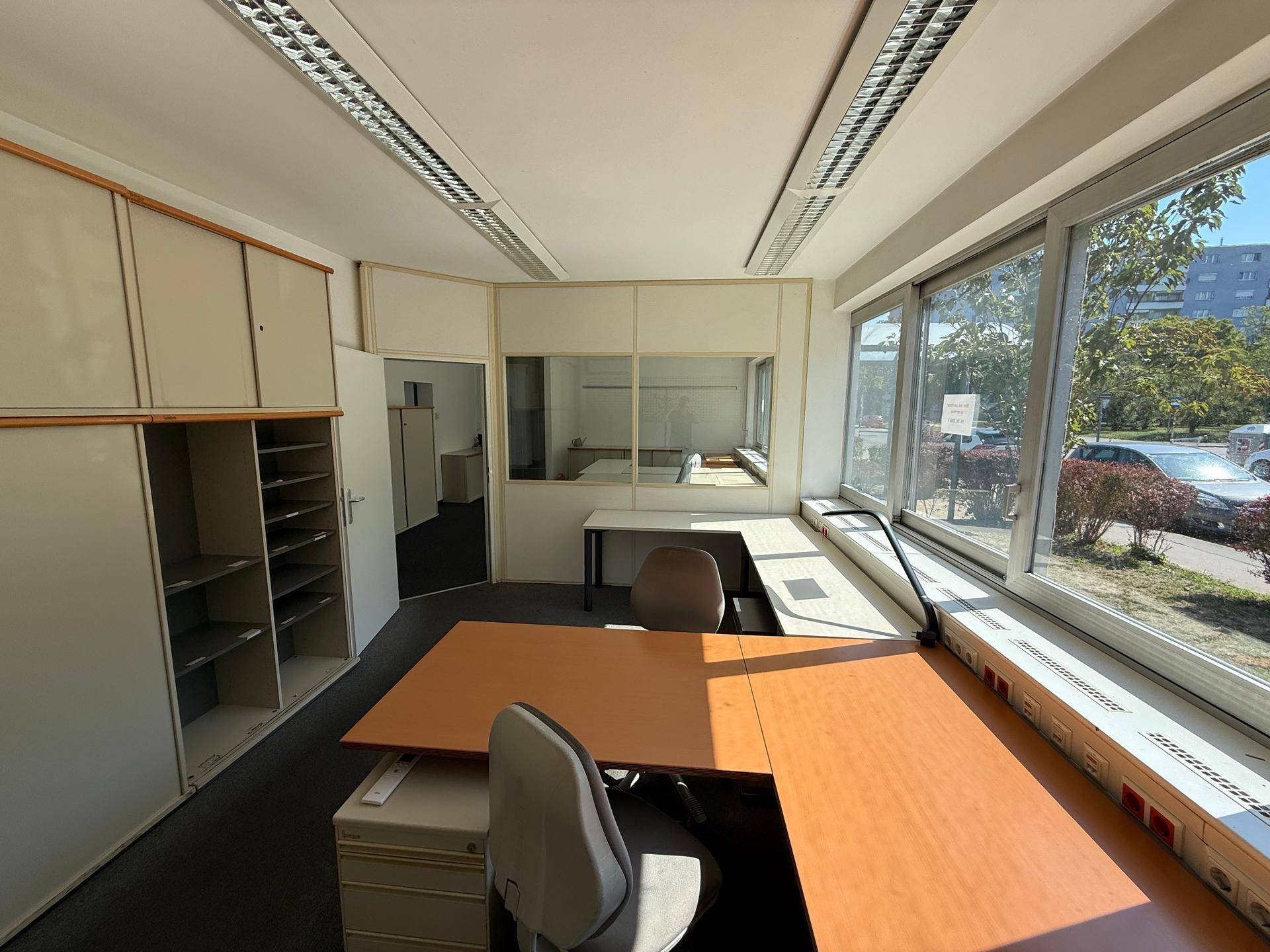 Helles Eckbüro mit Teppichboden, Schreibtischen und großen Fenstern mit Blick nach draußen.