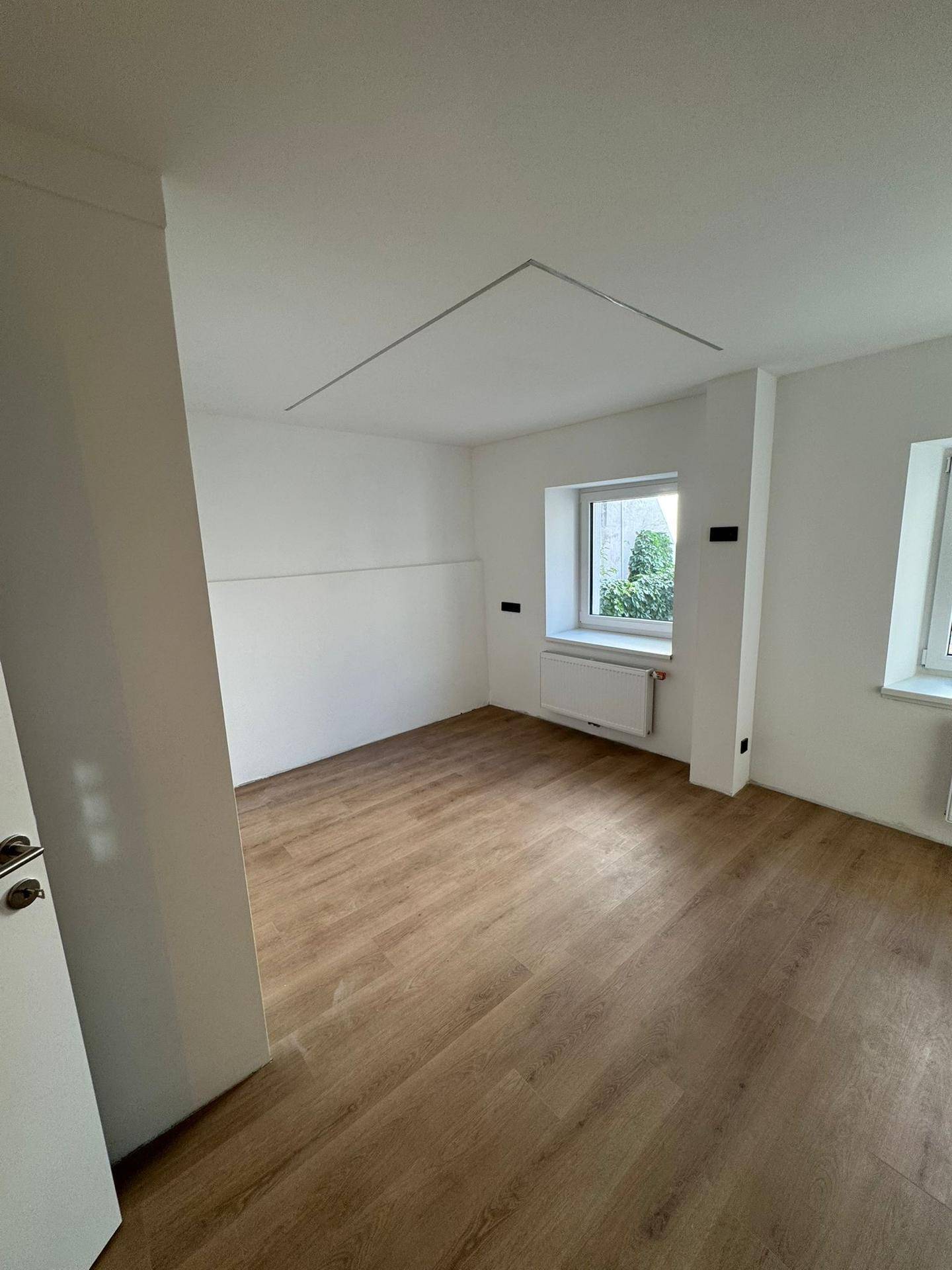 Helles, leeres Zimmer mit Laminatboden, weißen Wänden und einem Fenster mit Blick ins Grüne.