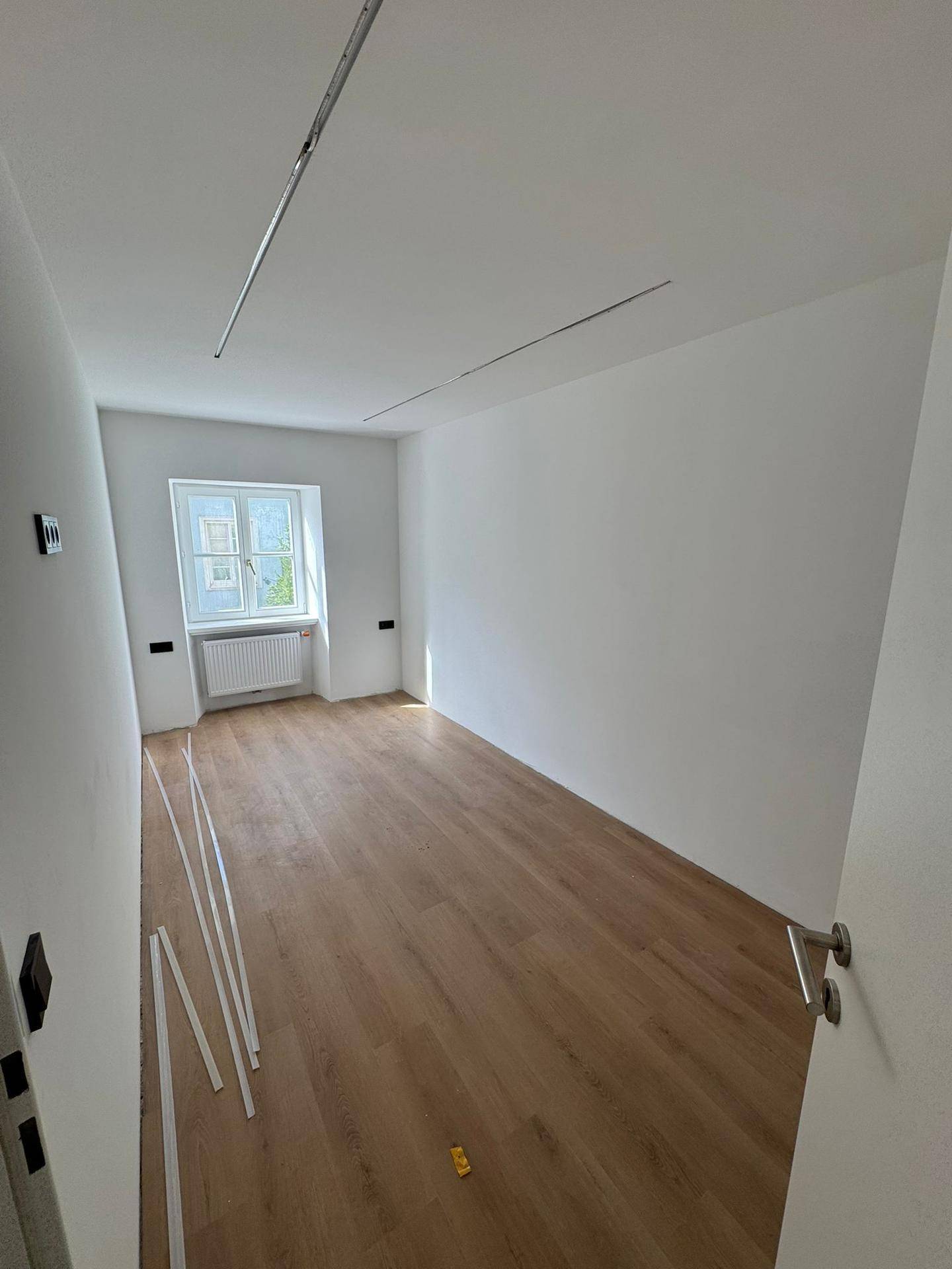 Geräumiges, helles Zimmer mit Laminatboden, weißen Wänden und einem großen Fenster.