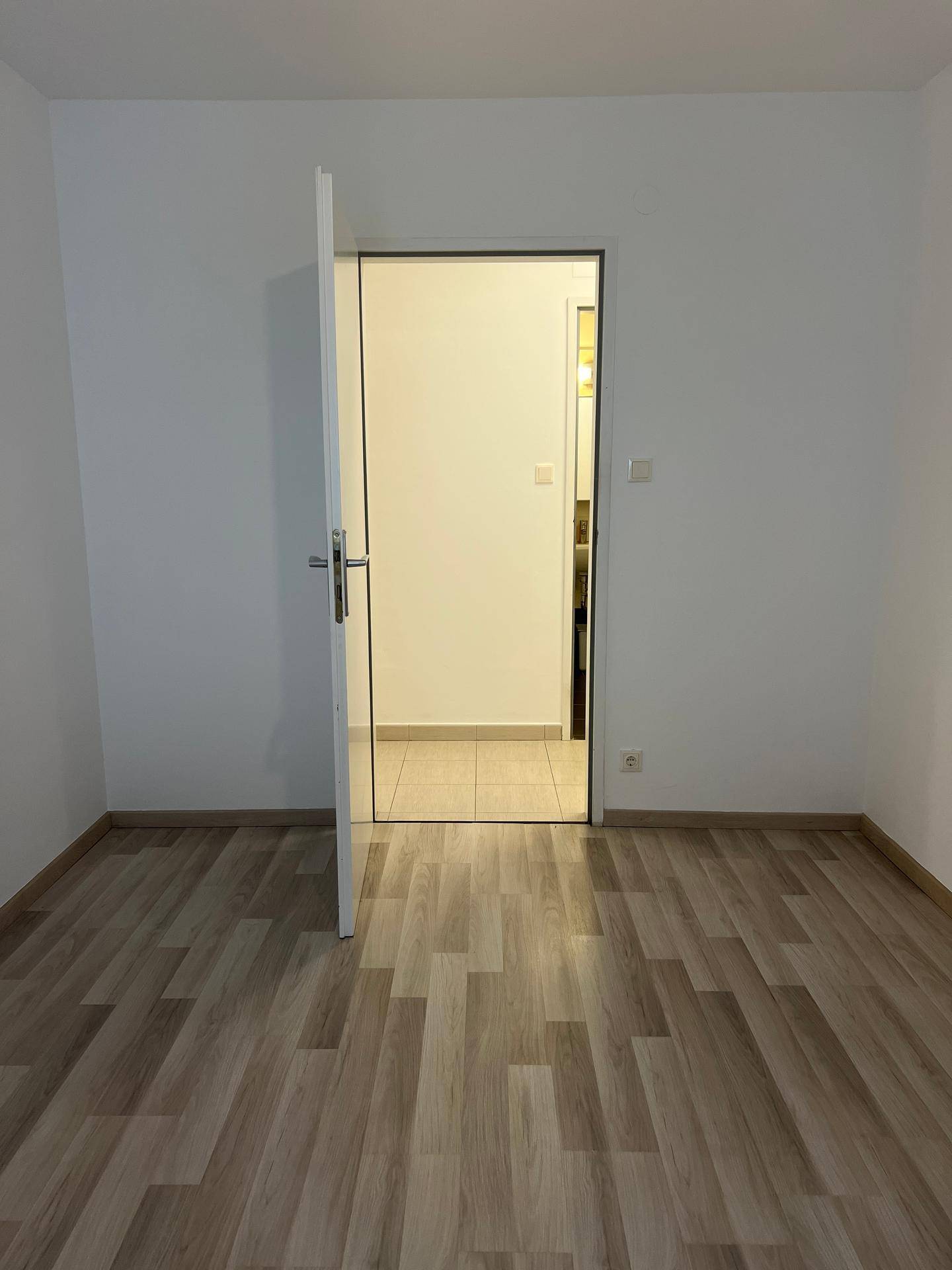Leeres Zimmer mit hellem Laminatboden und offener Tür, die zu einem gefliesten Bereich führt.