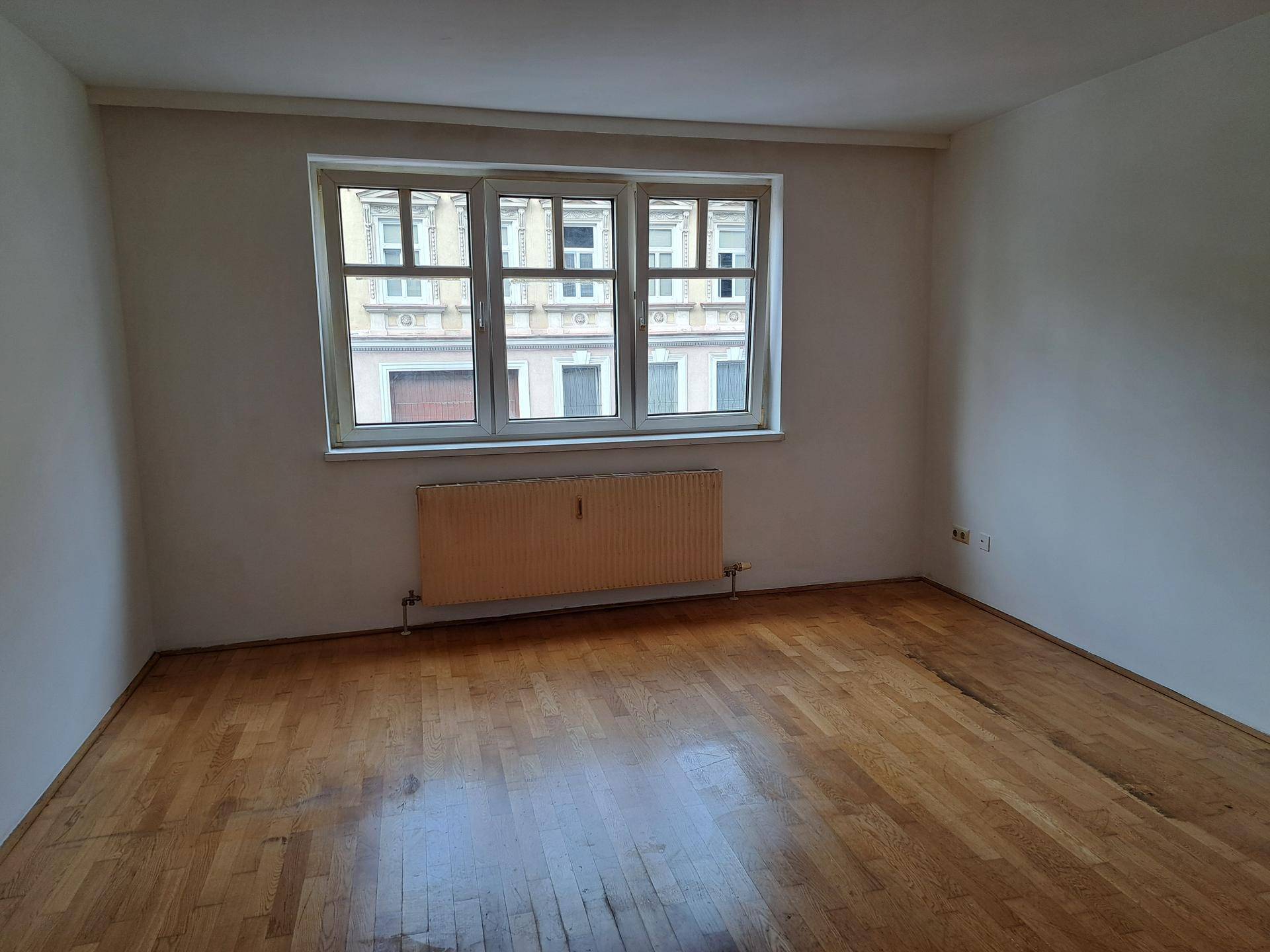 Helles Zimmer mit Holzboden, großem Fenster und Blick auf die gegenüberliegende Fassade.