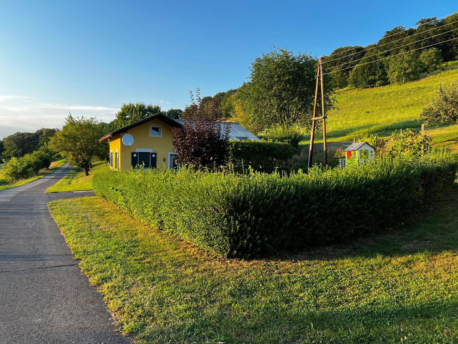 Weitläufige Ansicht des gelben Hauses mit Garten, Hecke und Spielhaus in grüner Hügellandschaft.