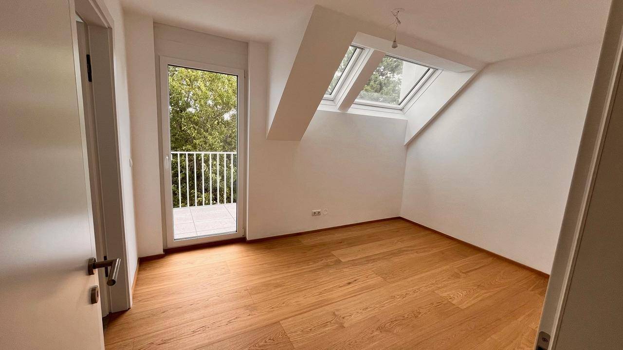 Schlafzimmer mit Holzboden, Dachfenster und Zugang zu einem kleinen Balkon mit Grünblick.