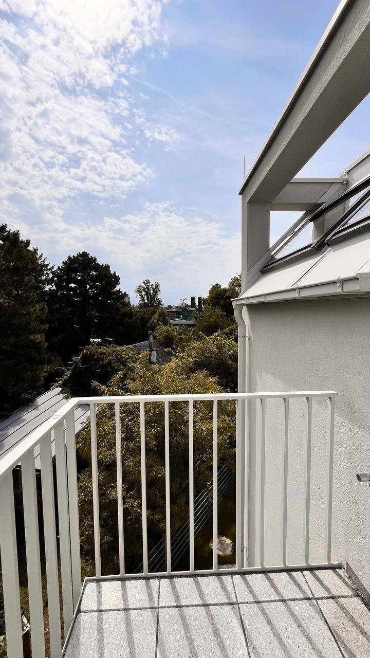 Kleiner Balkon mit Fliesenboden und Blick auf grüne Umgebung und Dächer unter blauem Himmel.