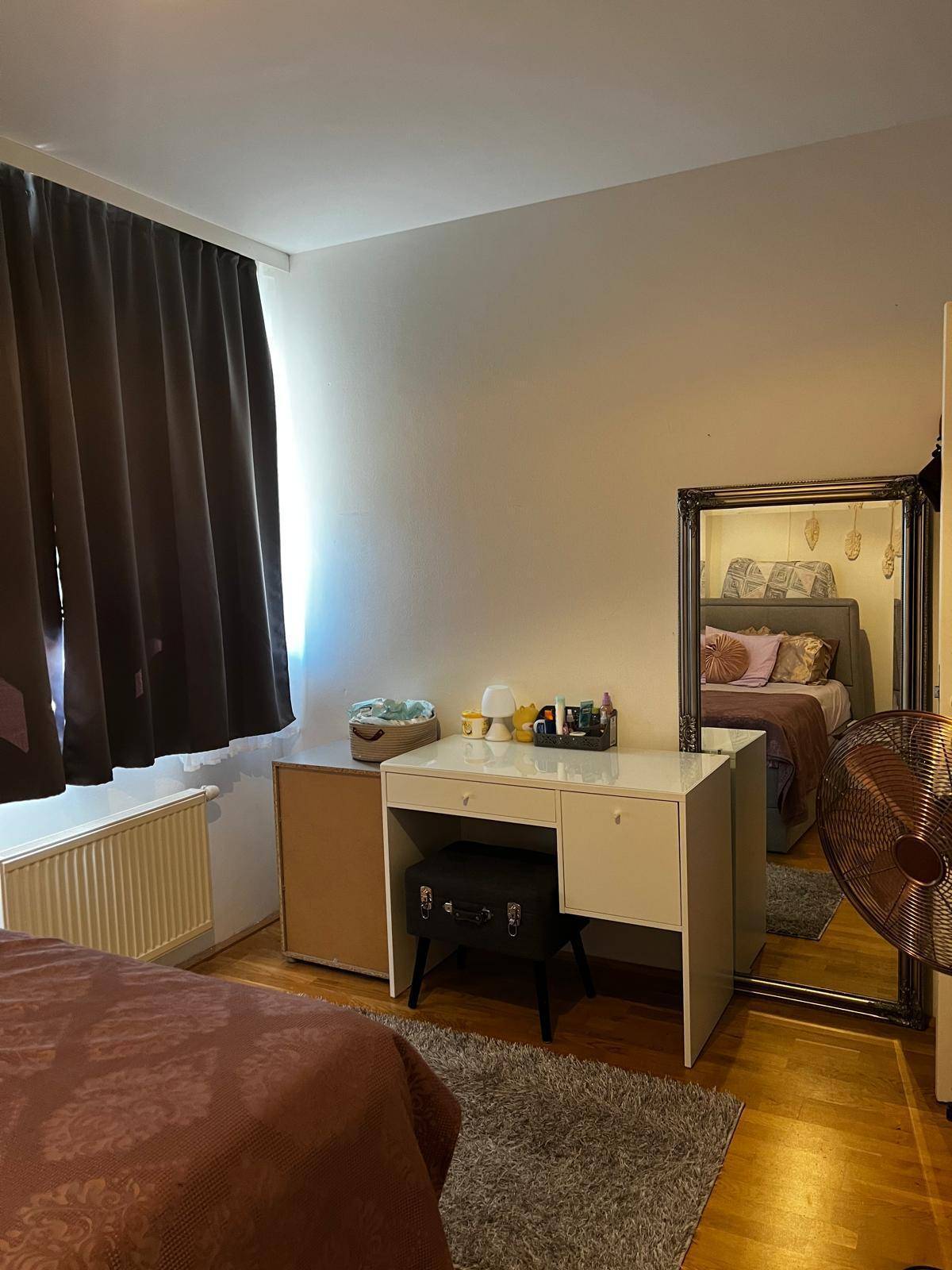 Schlafzimmer mit Bett, Schminktisch und großem Spiegel, beleuchtet durch ein Fenster mit dunklen Vorhängen.