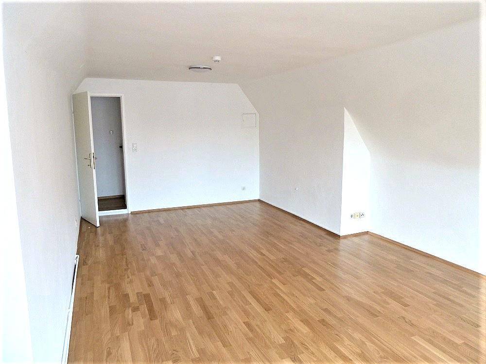 Geräumiger Dachgeschossraum mit Holzfußboden, weißen Wänden und Zugang zu einem weiteren Bereich.