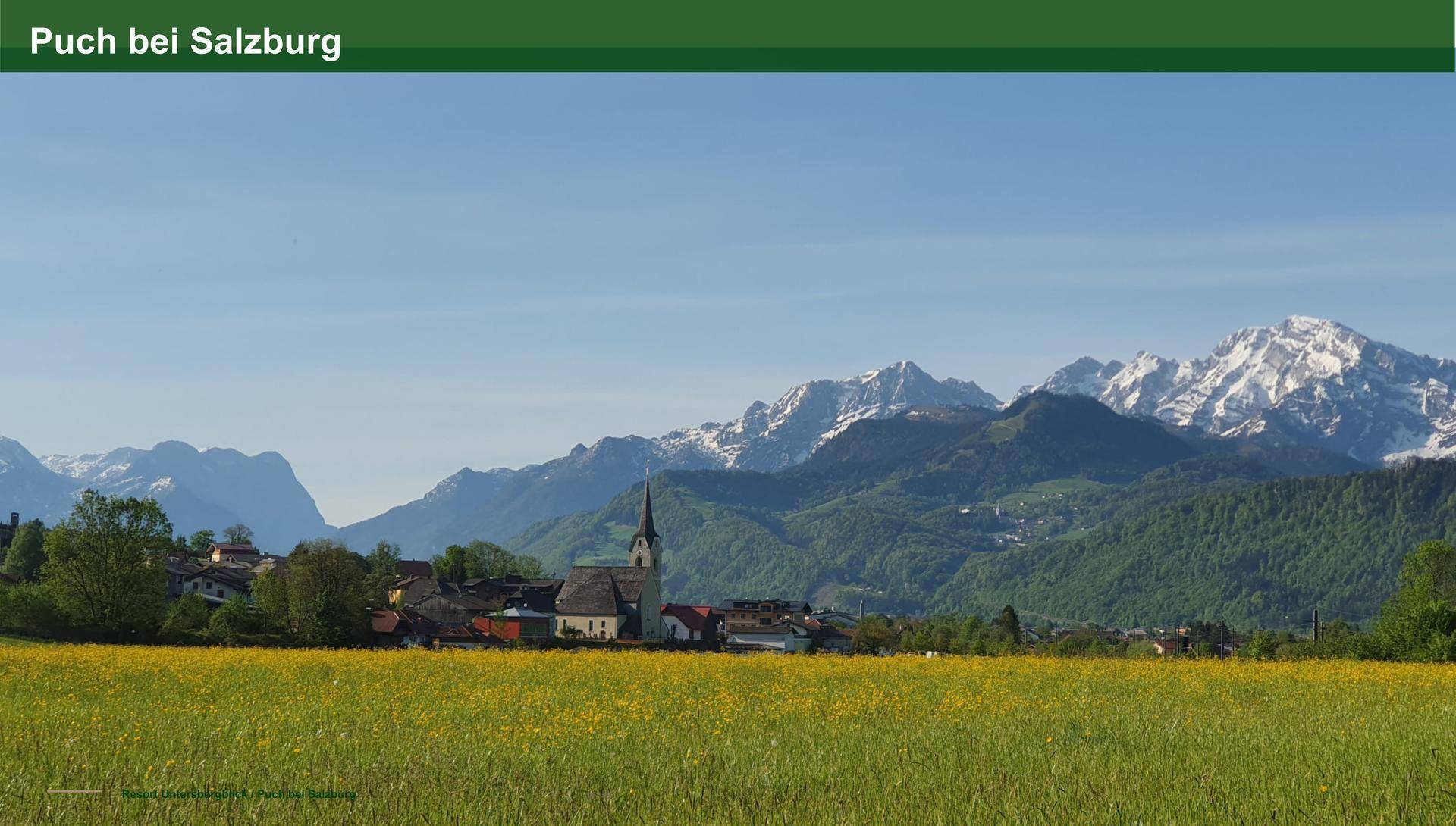Idyllische Landschaft mit einem Dorf, grünen Feldern und schneebedeckten Bergen im Hintergrund.