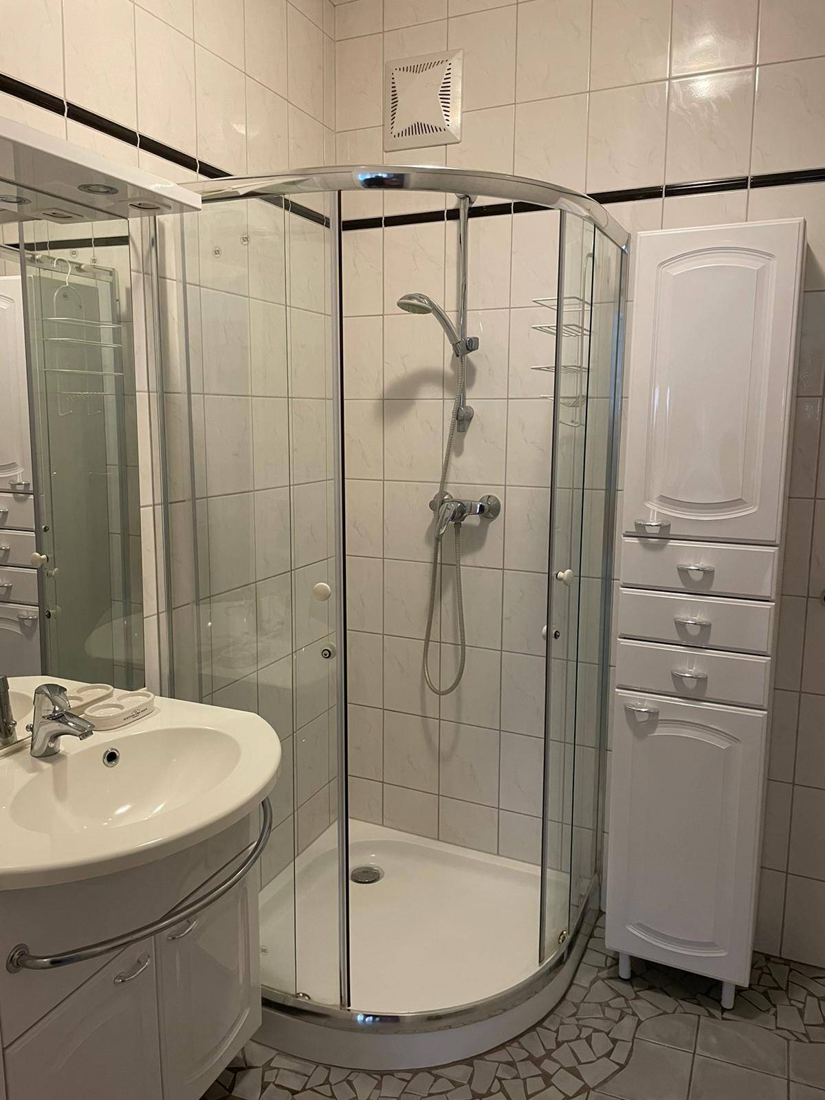 Badezimmer mit Eckdusche, Waschbecken und weißem Hochschrank, ausgestattet mit Fliesenboden.