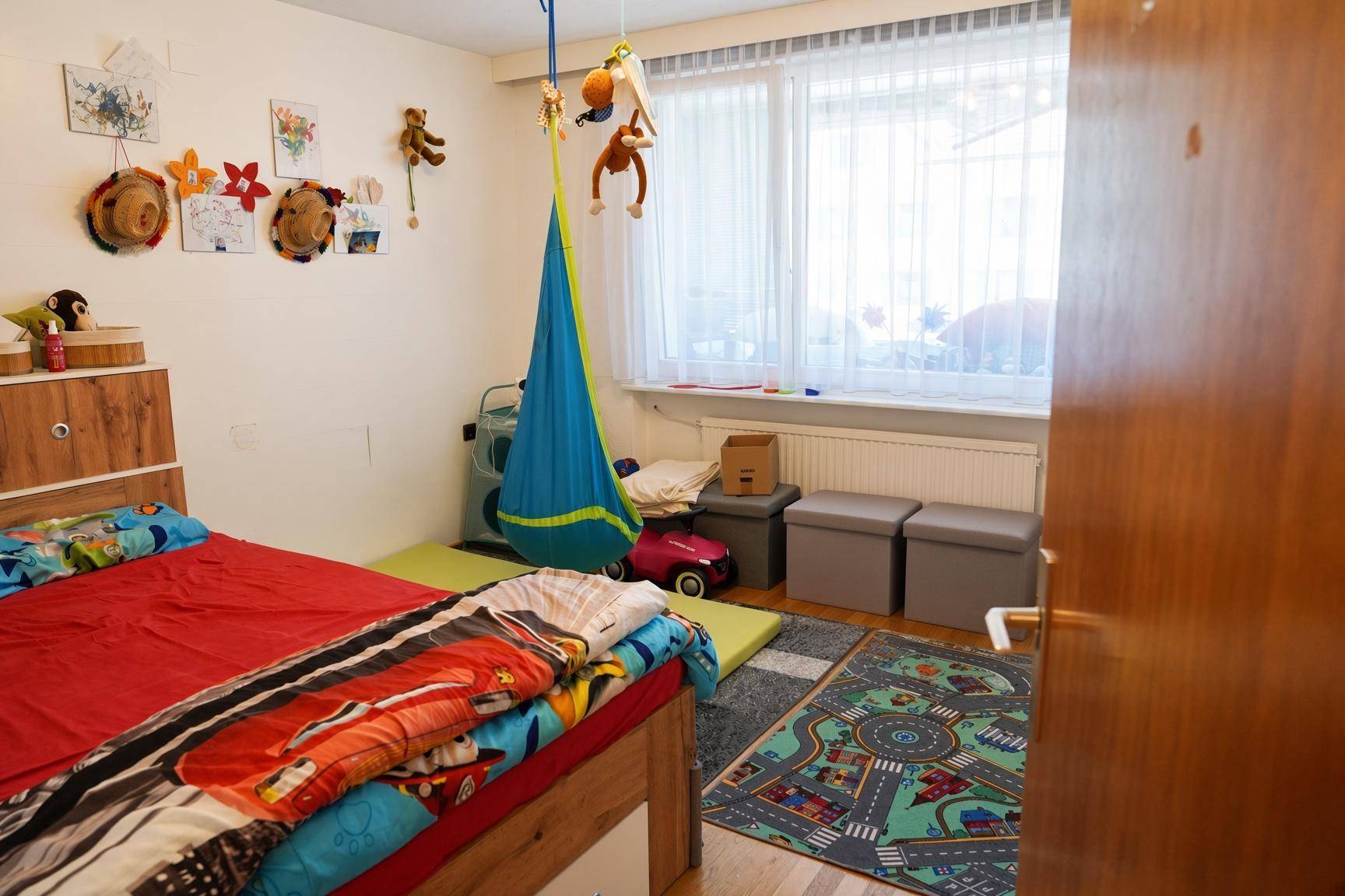 Kinderzimmer mit großem Fenster, Spielteppich und bunter Dekoration.