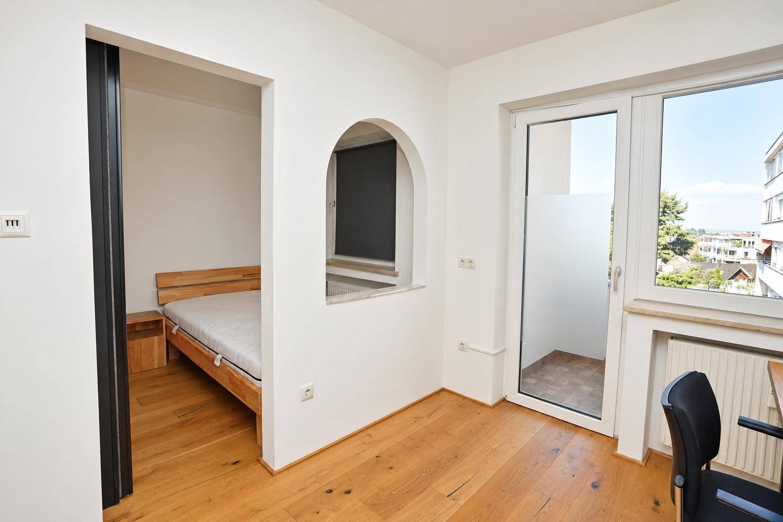 Offener Wohnbereich mit Holzboden und Durchgang zum Schlafzimmer, mit Zugang zu einem kleinen Balkon.