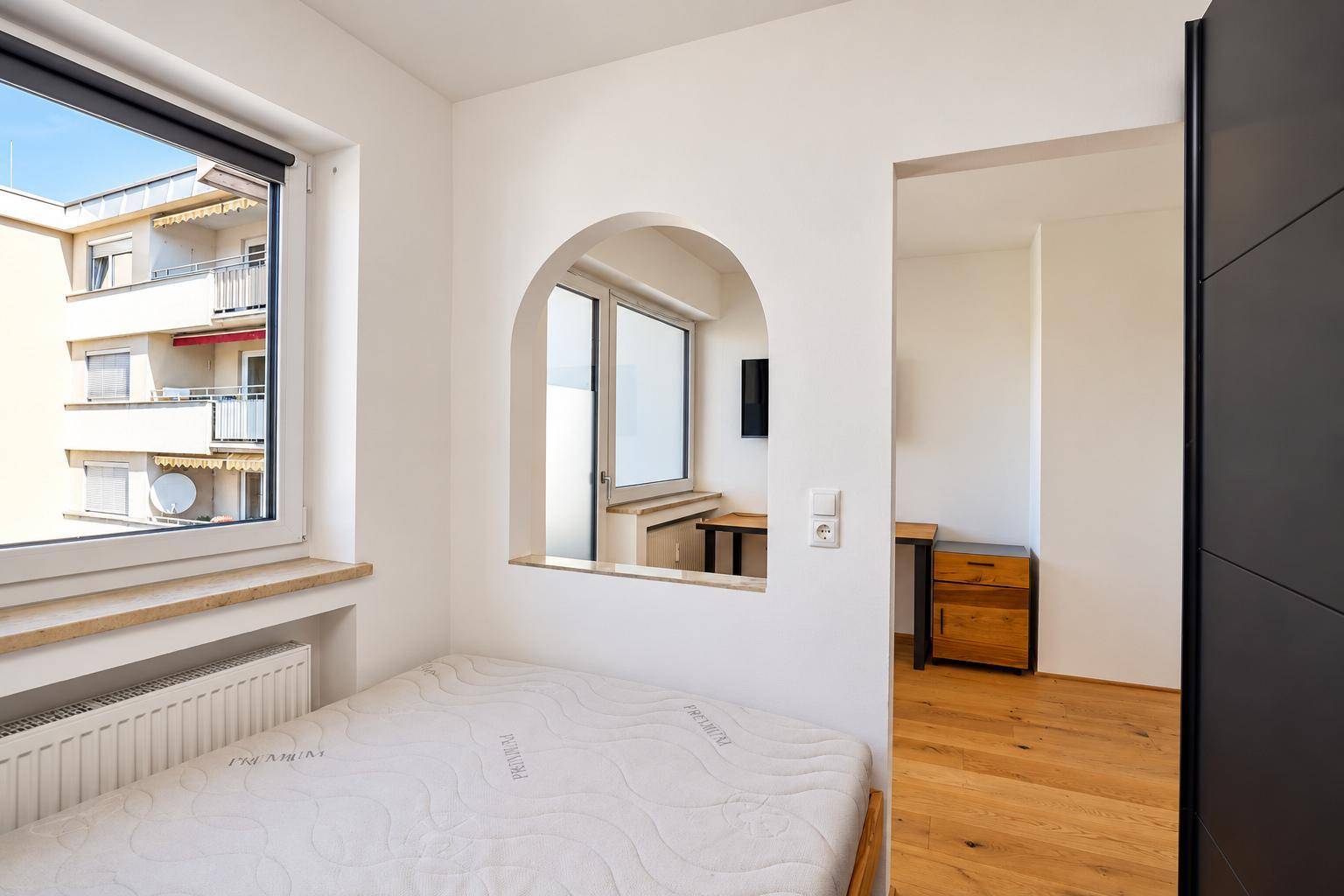 Schlafzimmer mit Holzbett und Fenster, das einen Blick in den angrenzenden Wohnbereich bietet.