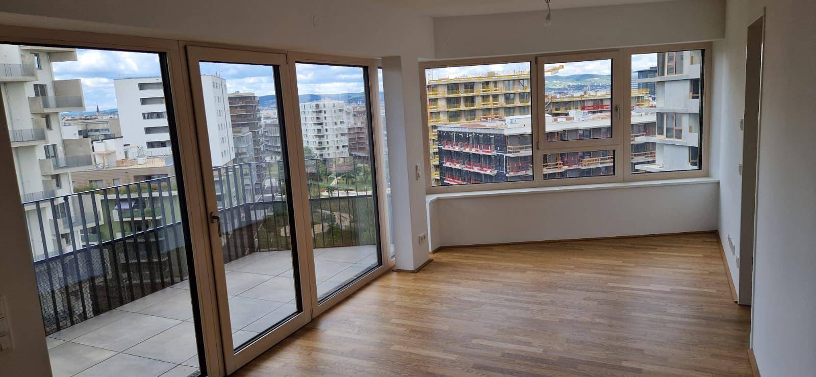 Geräumiger, heller Raum mit Holzboden und großen Fenstern mit Zugang zum Balkon und Stadtblick.