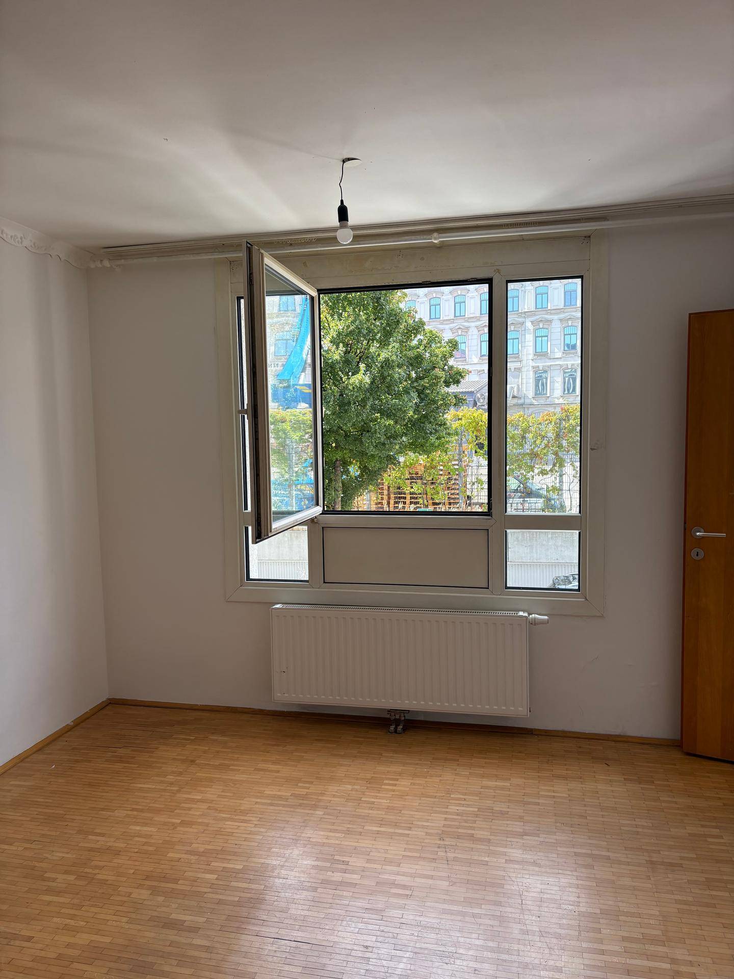 Heller Raum mit geöffnetem Fenster, Blick ins Grüne und auf die Stadt, ausgestattet mit Parkettboden.