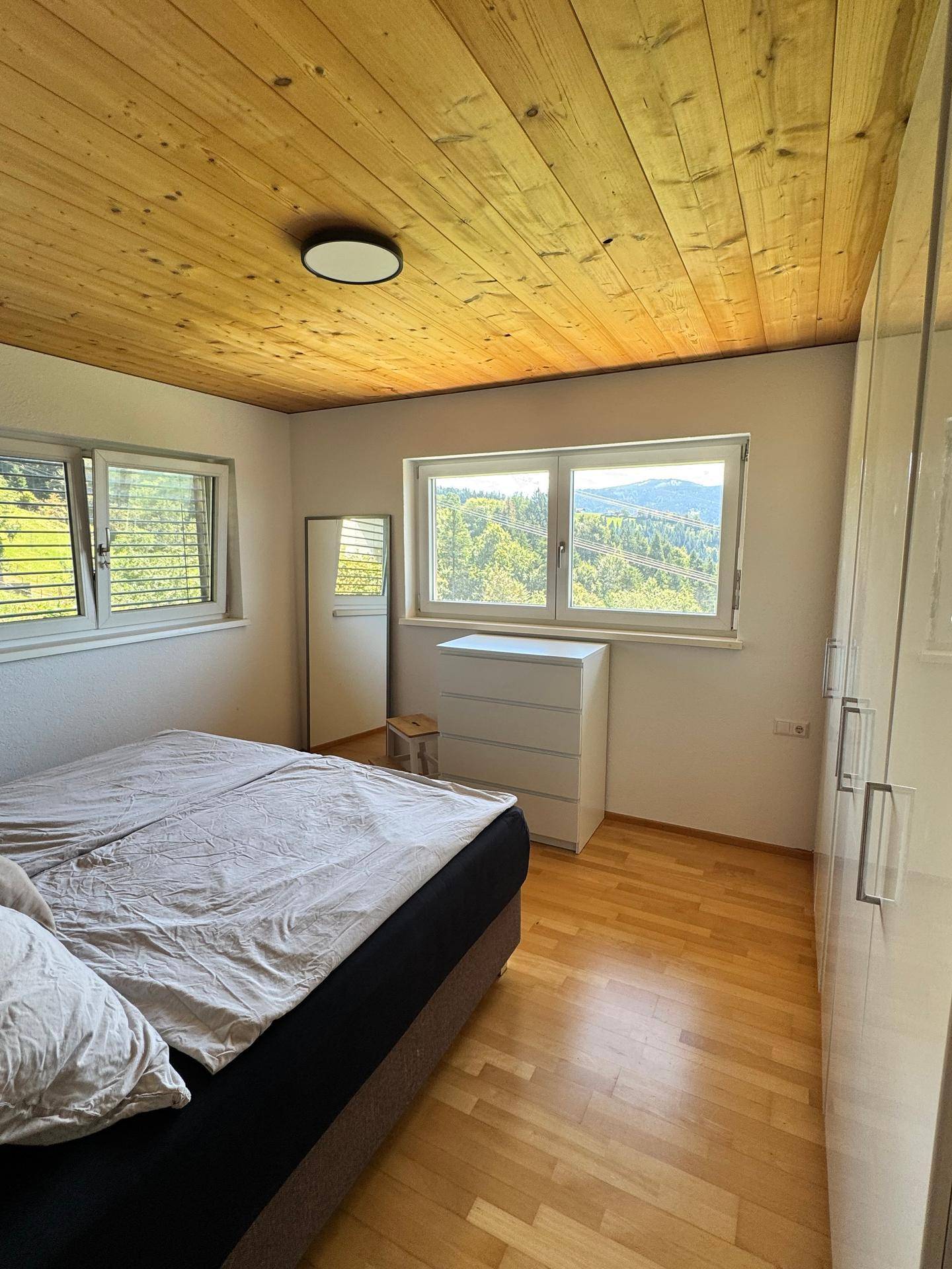 Helles Schlafzimmer mit Holzboden, weißen Schränken und Fenstern mit Blick auf die grüne Landschaft.