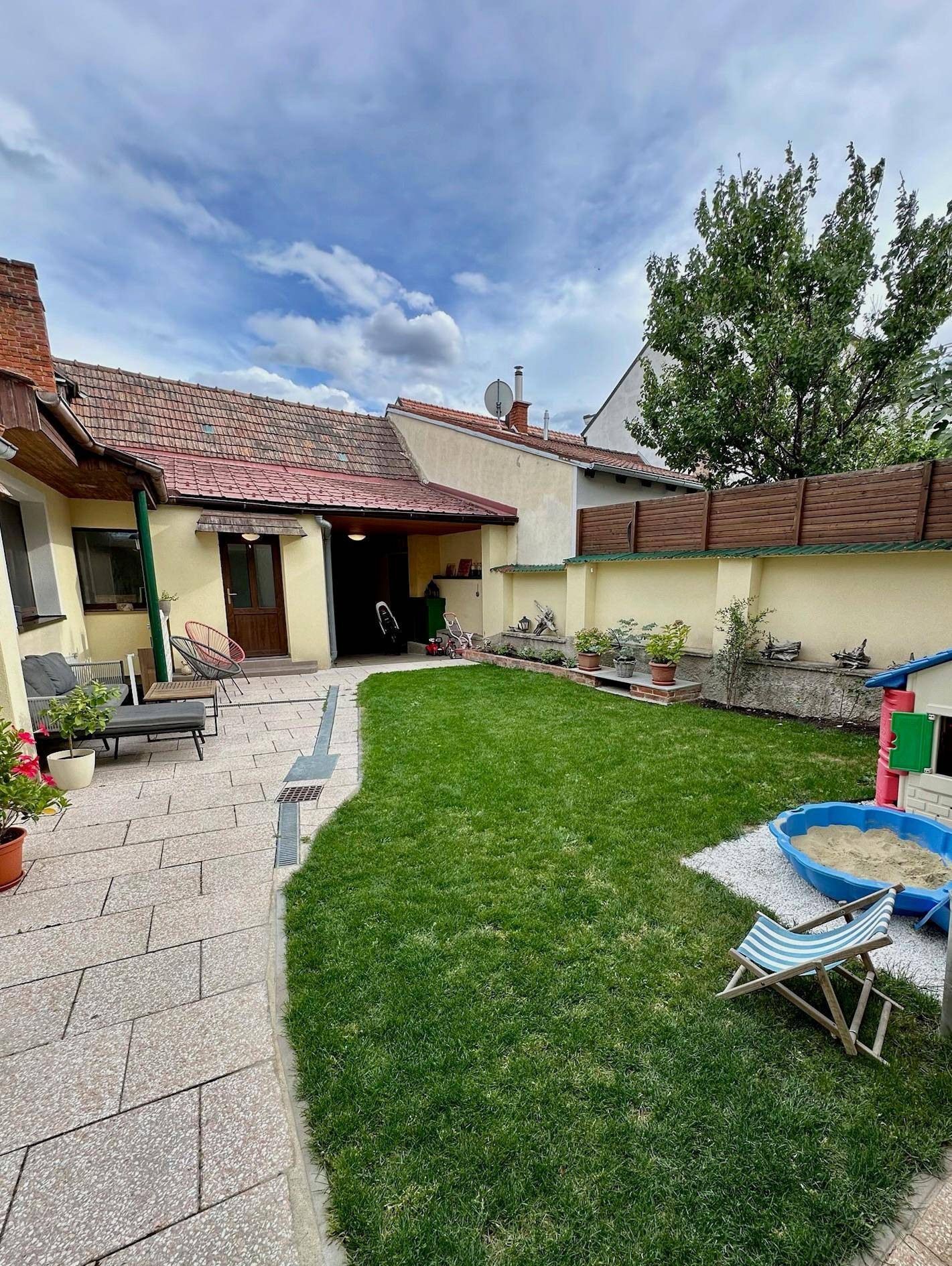 Gepflegter Garten mit Rasenfläche, gepflasterter Terrasse und Kinderspielbereich.