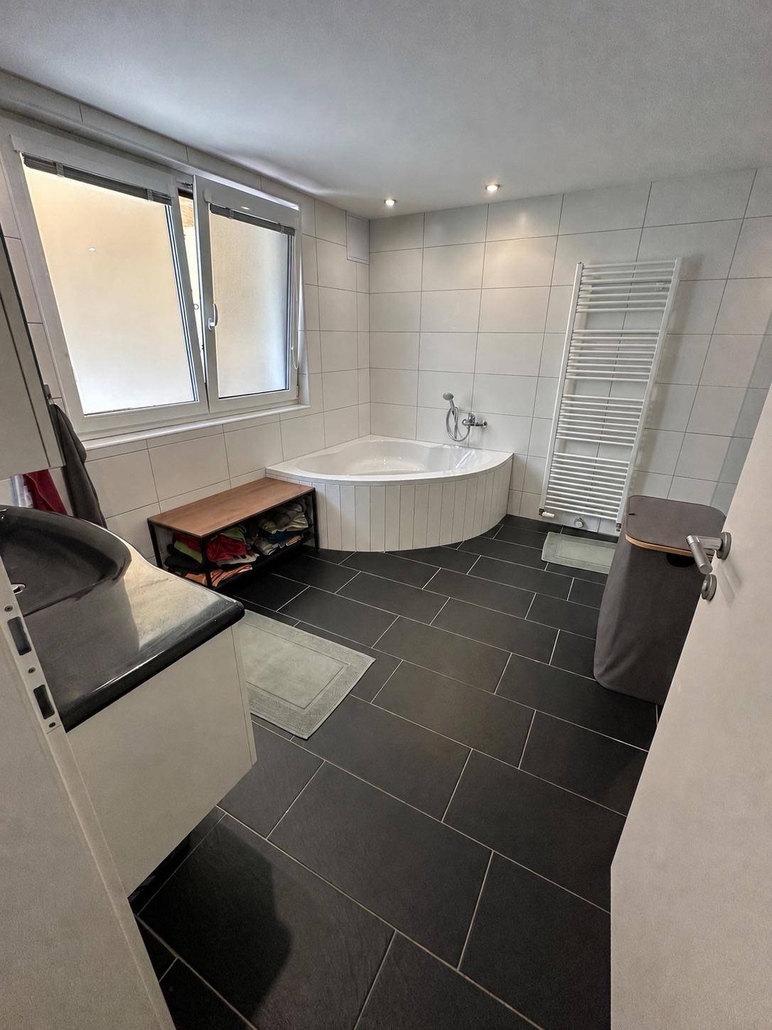 Badezimmer mit Eckbadewanne, dunklen Fliesen und Fenster.