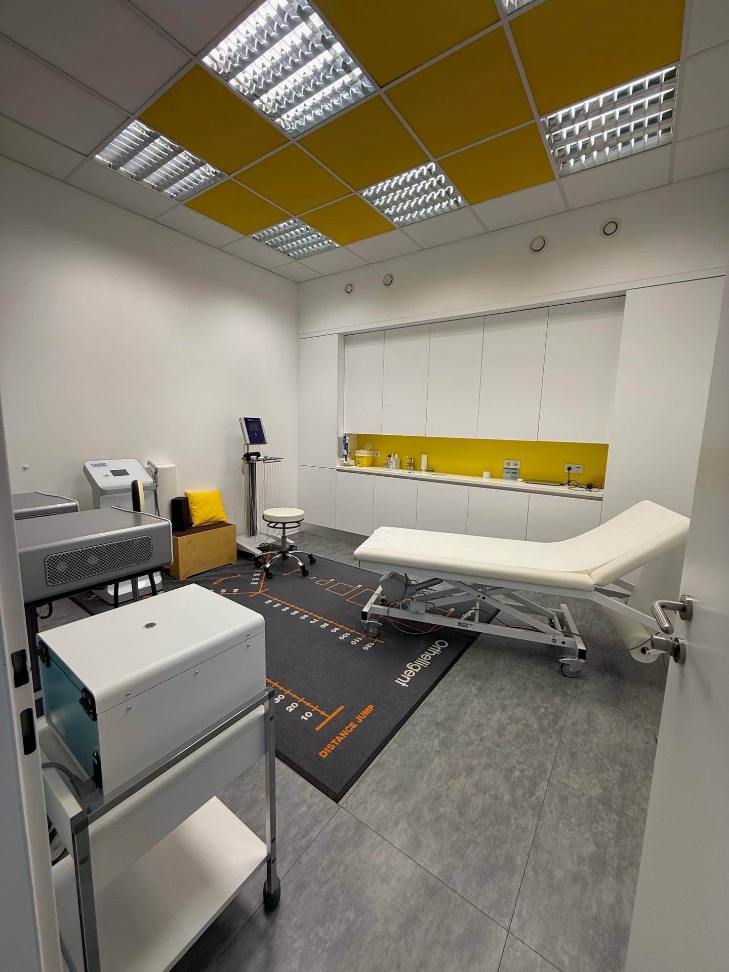 Behandlungsraum mit Liege, medizinischen Geräten und gelben Akzenten an der Decke.