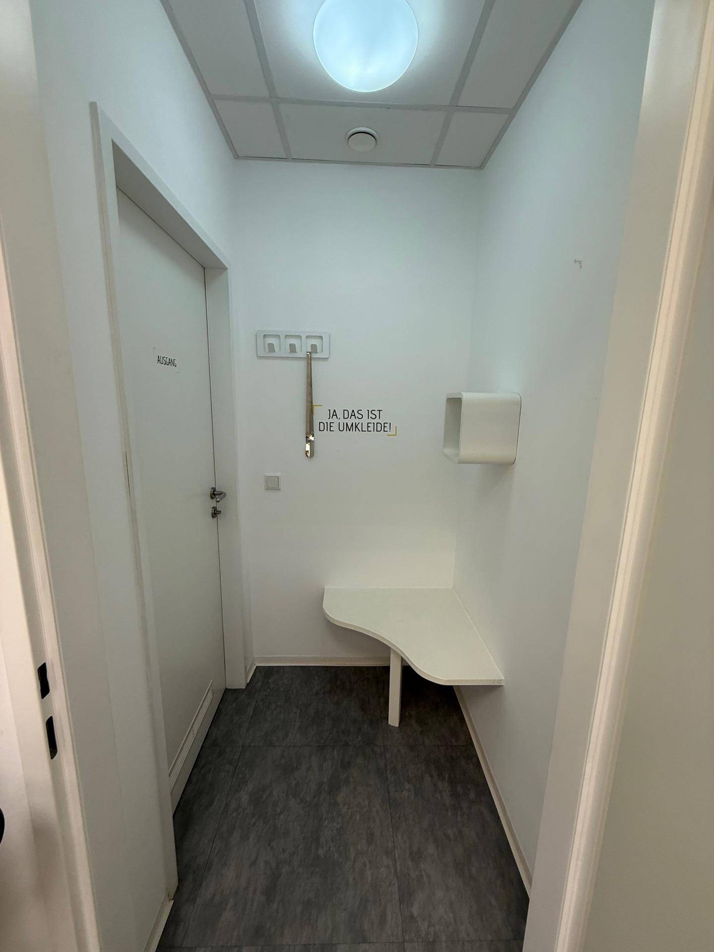 Kleine Umkleidekabine mit Sitzbank und Haken, funktional und schlicht in Weiß gehalten.
