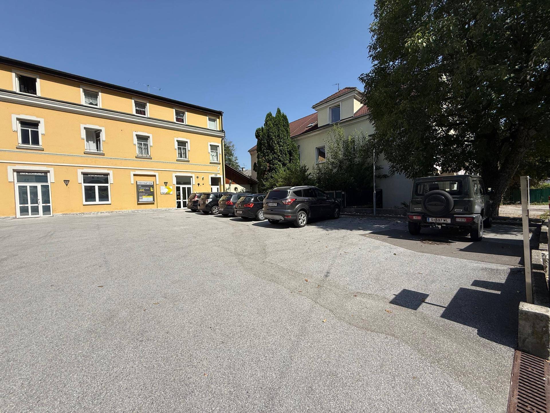 Großer asphaltierter Parkplatz vor einem gelben Gebäude mit weiteren Autos und Bäumen im Hintergrund.