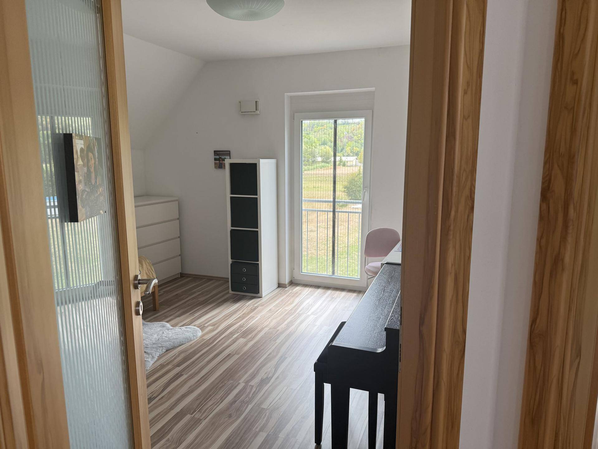 Freundliches Zimmer mit Holzboden, Regal und Balkontür, die viel Tageslicht hereinlässt.