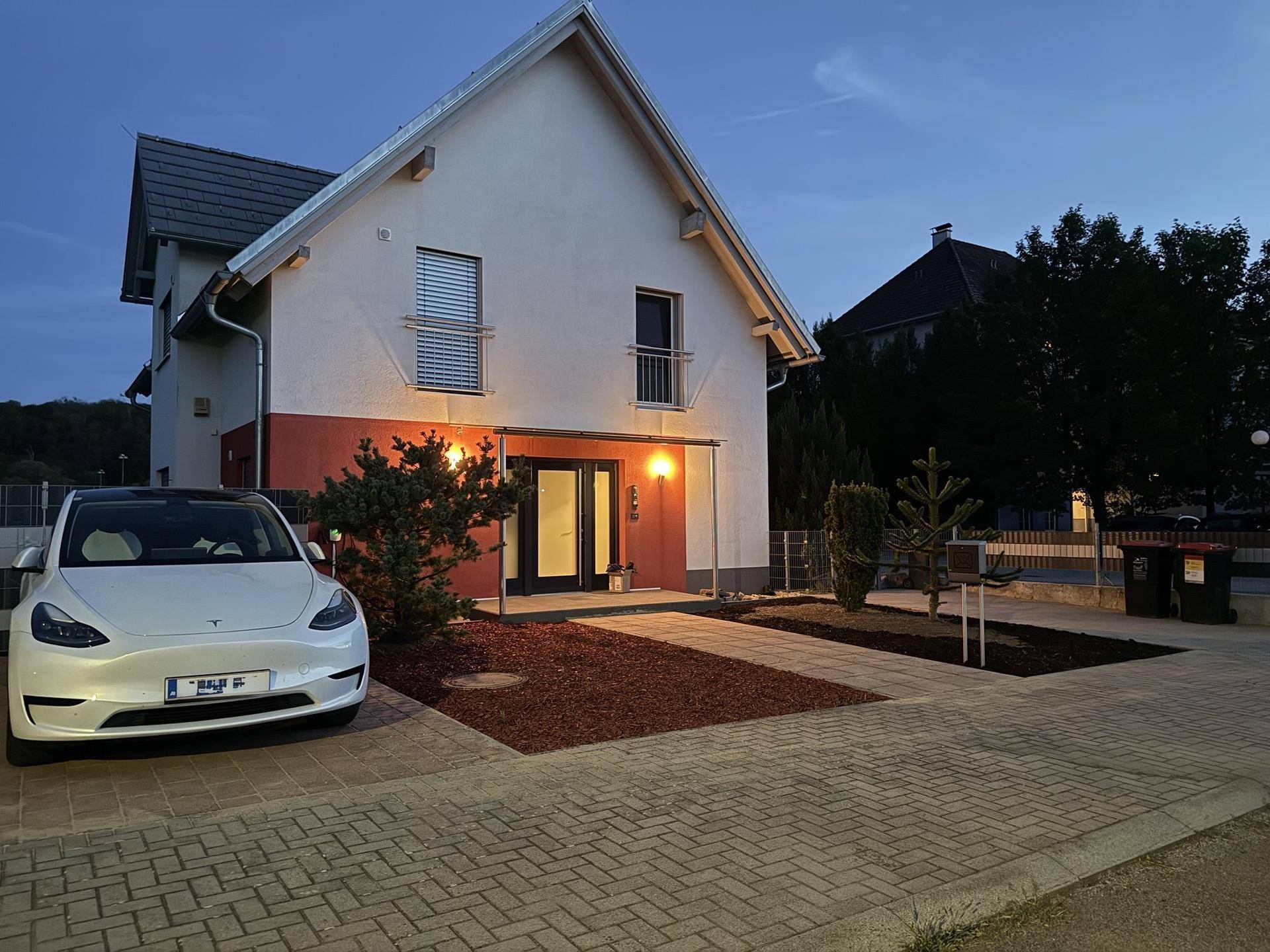 Das Einfamilienhaus bei Abenddämmerung mit beleuchteter Fassade und einem geparkten Elektrofahrzeug.