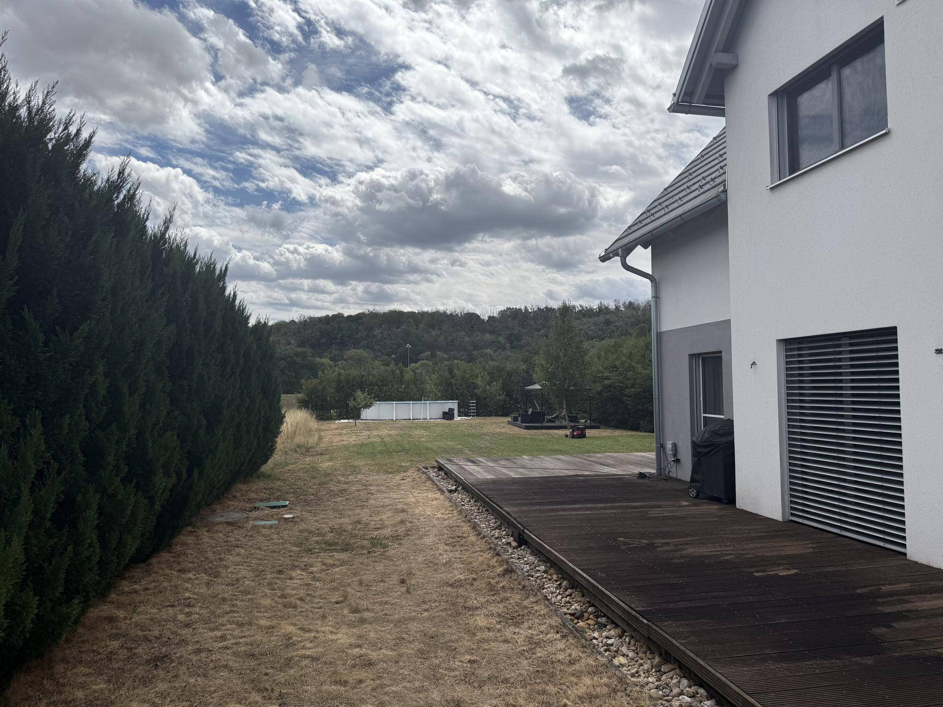 Großer Gartenbereich mit Holzterrasse, umgeben von hohen Hecken und Bäumen.