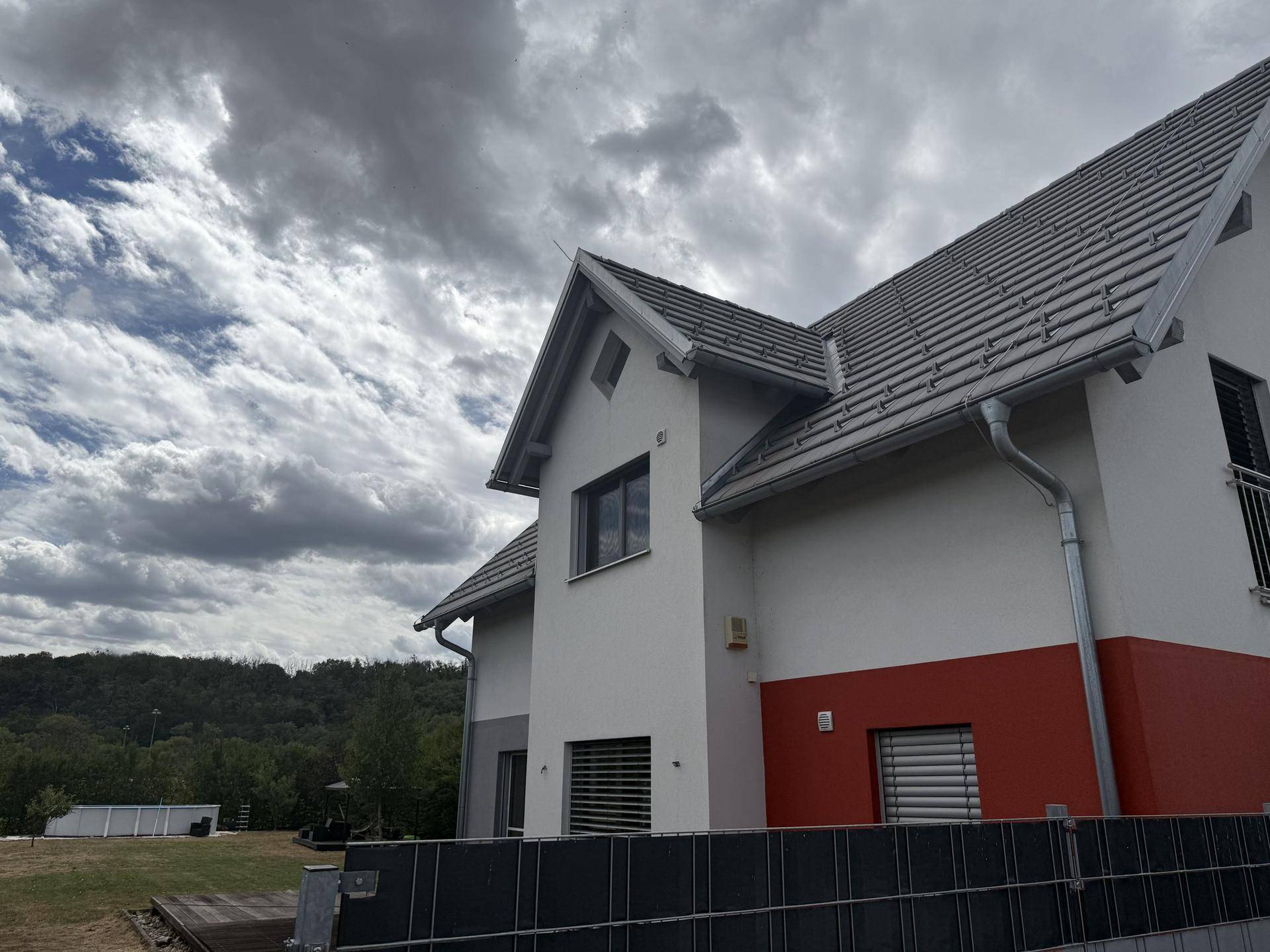Seitenansicht des Hauses mit Satteldach, weißer und roter Fassade unter bewölktem Himmel.