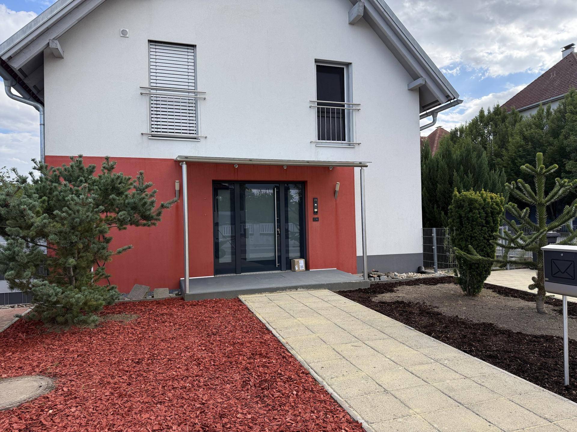 Eingangsbereich des Hauses mit roter Fassade, überdachtem Eingang und gepflastertem Zugangsweg.