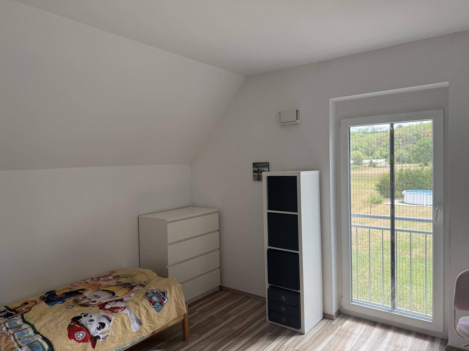 Helles Zimmer mit Kommode, Regal und Blick auf den Garten mit kleinem Pool.