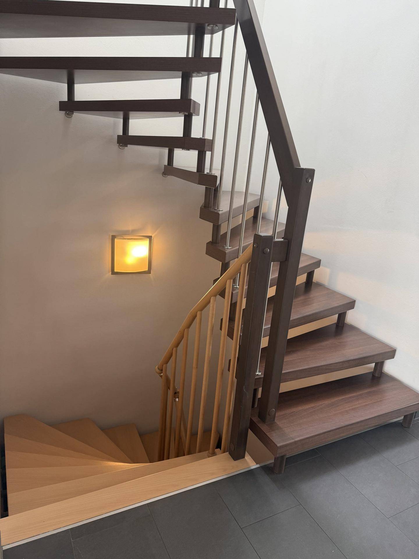 Holztreppe mit Metallgeländer, die in die obere Etage führt, beleuchtet von einer Wandleuchte.