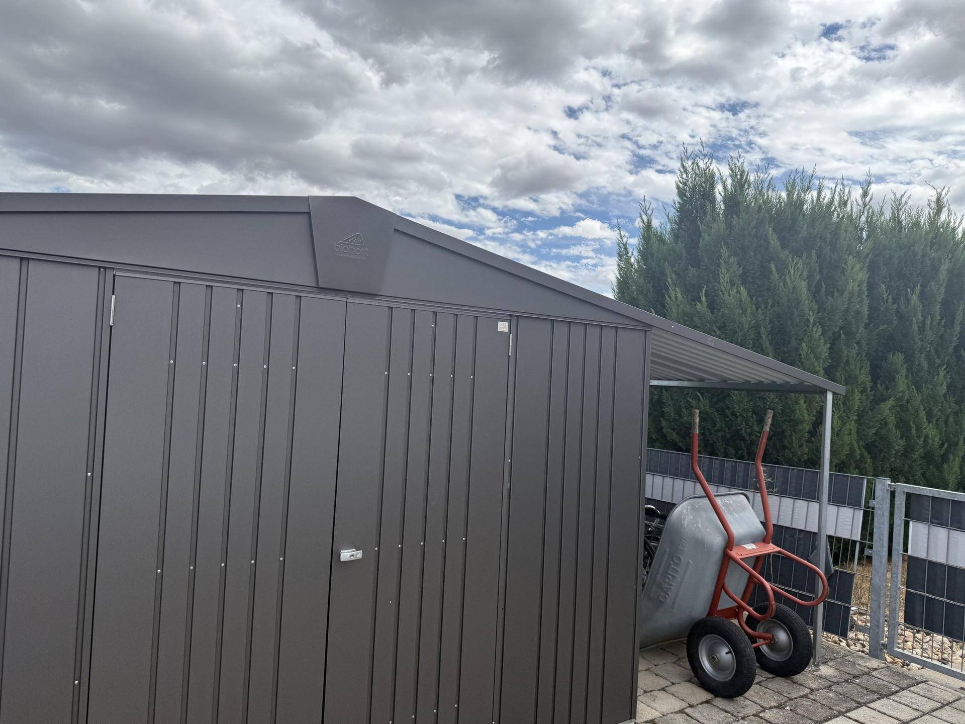 Moderner Gartenschuppen aus Metall mit Vordach und einer Schubkarre im Außenbereich.