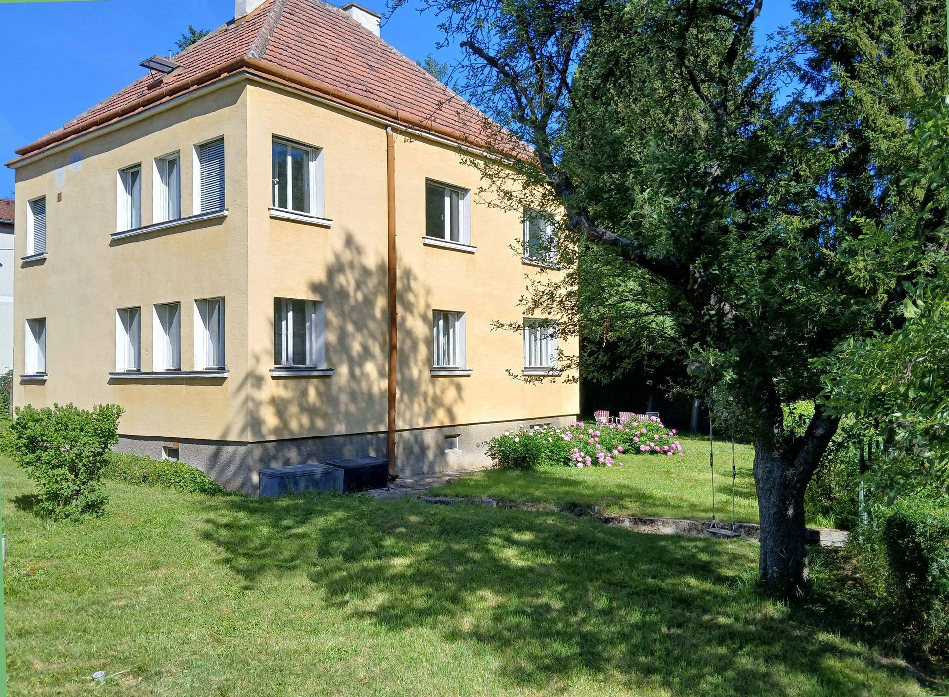Gelbes Wohnhaus mit rotem Ziegeldach, umgeben von einem grünen Garten und Bäumen.