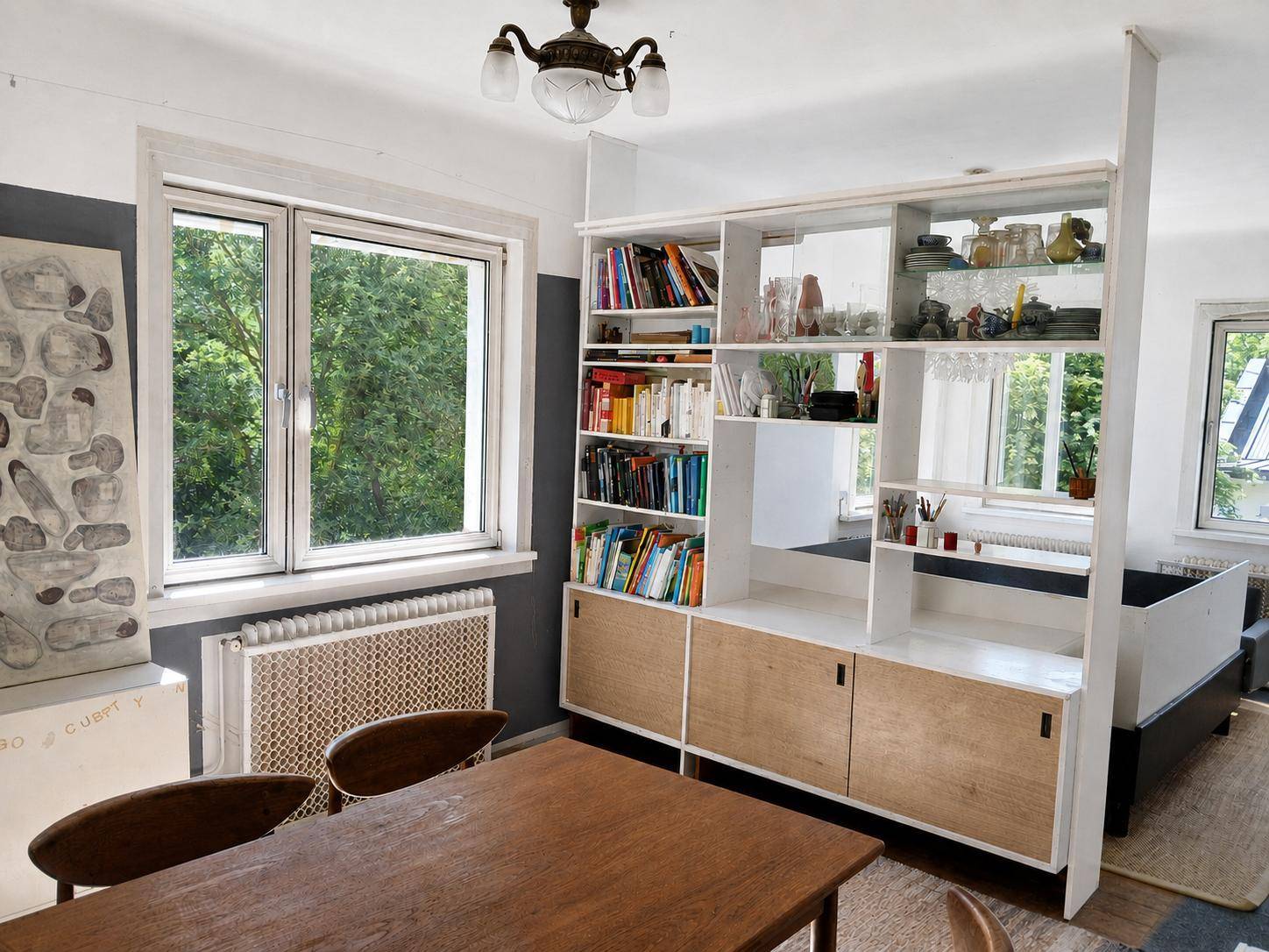 Offener Wohn-Essbereich mit Holztisch, Bücherregal als Raumteiler und großen Fenstern.