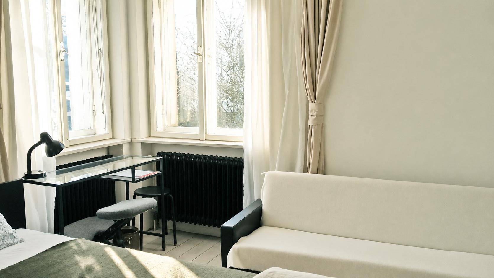Helles Schlafzimmer mit Bett, Sofa, Schreibtisch und großen Fenstern mit Vorhängen.
