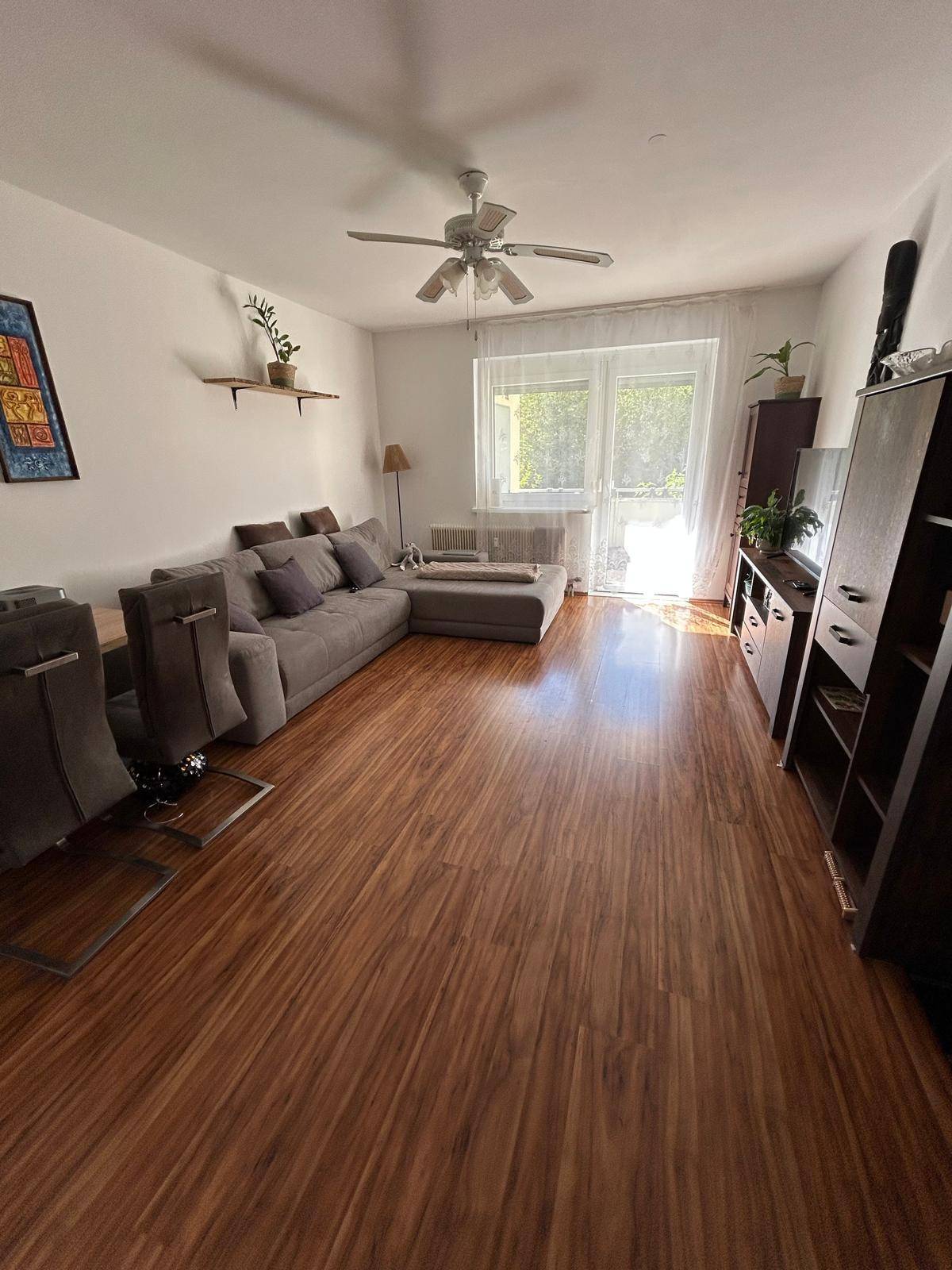 Helles Wohnzimmer mit großem Ecksofa, Laminatboden und Deckenventilator, gut beleuchtet durch ein großes Fenster.